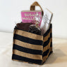 Gift basket from Saratoga Chocolate Co. displayed on a white background, featuring a burlap tote bag with black stripes filled with the book “Sweet Success” by Susan Mallery, Fleur de Lait chocolate bar, Dark Chocolate Covered Almonds, and a Milk Chocolate Peanut Butter Cup.