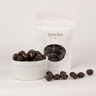 Image shows a pile of dark chocolate coated espresso beans displayed in a fluted white bowl with pile of dark chocolate espresso beans scattered in front. Behind the bowl and offset to the right is a pouch of dark chocolate covered espresso beans representing how the product is packaged.