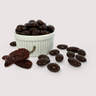 Image shows a pile of dark chocolate coated chipotle spiced almonds displayed in a fluted white bowl with some dark chocolate coated chipotle spiced almonds and two chipotle chilis scattered in front of the bowl.