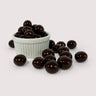 Image shows a pile of dark chocolate coated macadamia nuts displayed in a fluted white bowl with some dark chocolate coated macadamia nuts scattered in front