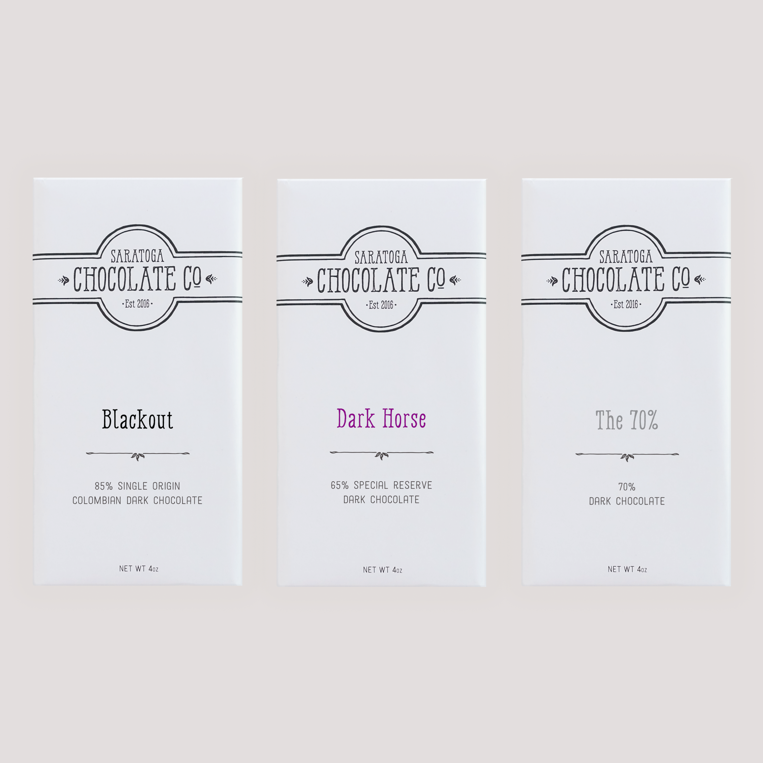 Three Saratoga Chocolate Co. dark chocolate bars in white wrappers with colorful text: Blackout, Dark Horse and 70%, displayed side by side on a light background.