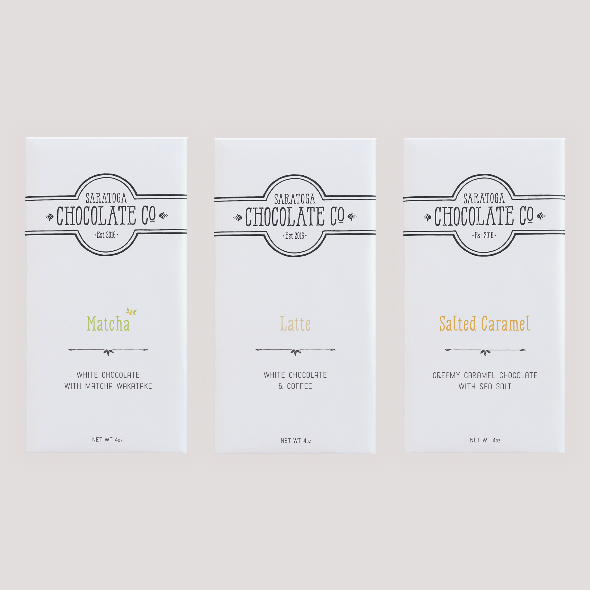 Three Saratoga Chocolate Co. white chocolate bars in white wrappers with colorful text: Matcha, Latte and Salted Caramel, displayed side by side on a light background.