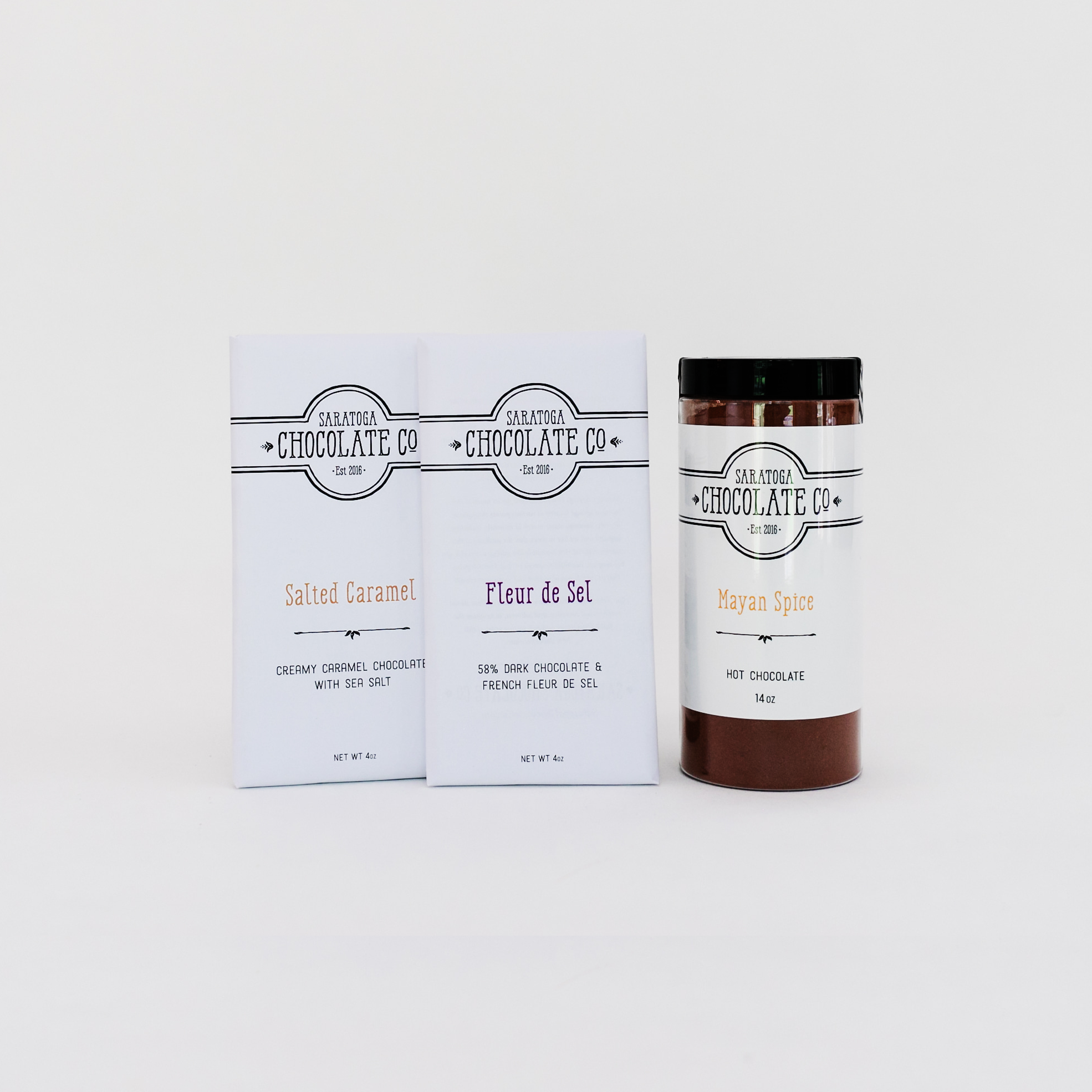 Saratoga Chocolate Co. Sugar & Spice Set featuring a 14-ounce jar of Mayan Spice Hot Chocolate Mix, labeled and sealed, alongside two 4-ounce chocolate bars—Fleur de Sel and Salted Caramel—wrapped in signature white paper with the Saratoga Chocolate Co. logo. The Mayan Spice Hot Chocolate is a bold dark chocolate blend infused with cinnamon, chili, and cayenne pepper. The Fleur de Sel Bar is dark chocolate with French sea salt, and the Salted Caramel Bar is creamy caramelized white chocolate with a touch of