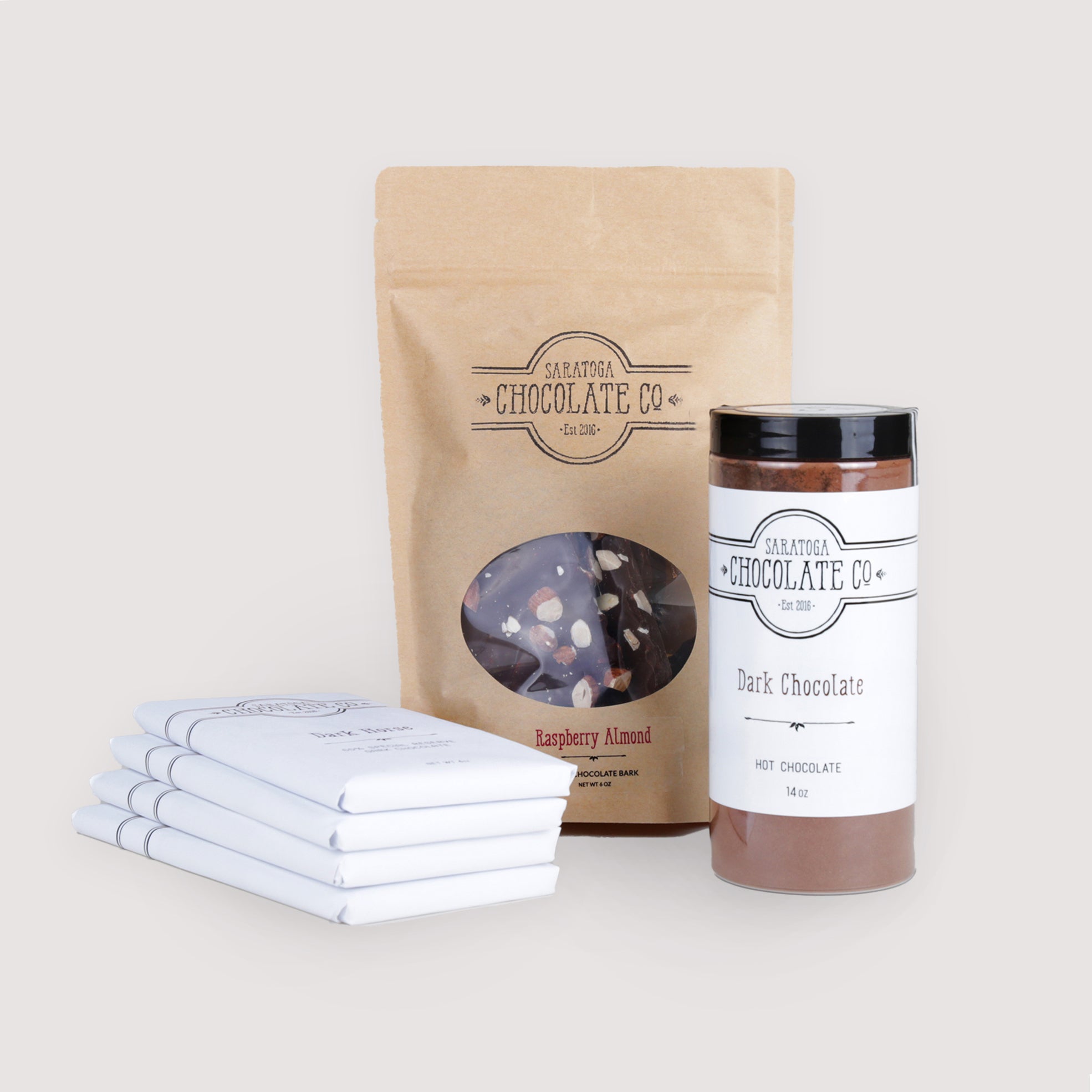 Saratoga Chocolate Co. Dark Chocolate Collection featuring four 4-ounce dark chocolate bars—Velvet, Dark Horse, The 70%, and Blackout—each wrapped in signature white packaging with branded labels. Includes a 6-ounce Raspberry Almond Dark Chocolate Bark in a kraft paper zipper-sealed bag with a clear window displaying raspberry and almond toppings, and a 14-ounce jar of Dark Hot Chocolate mix labeled and sealed. A dairy- and gluten-free artisanal chocolate gift set perfect for dark chocolate lovers.