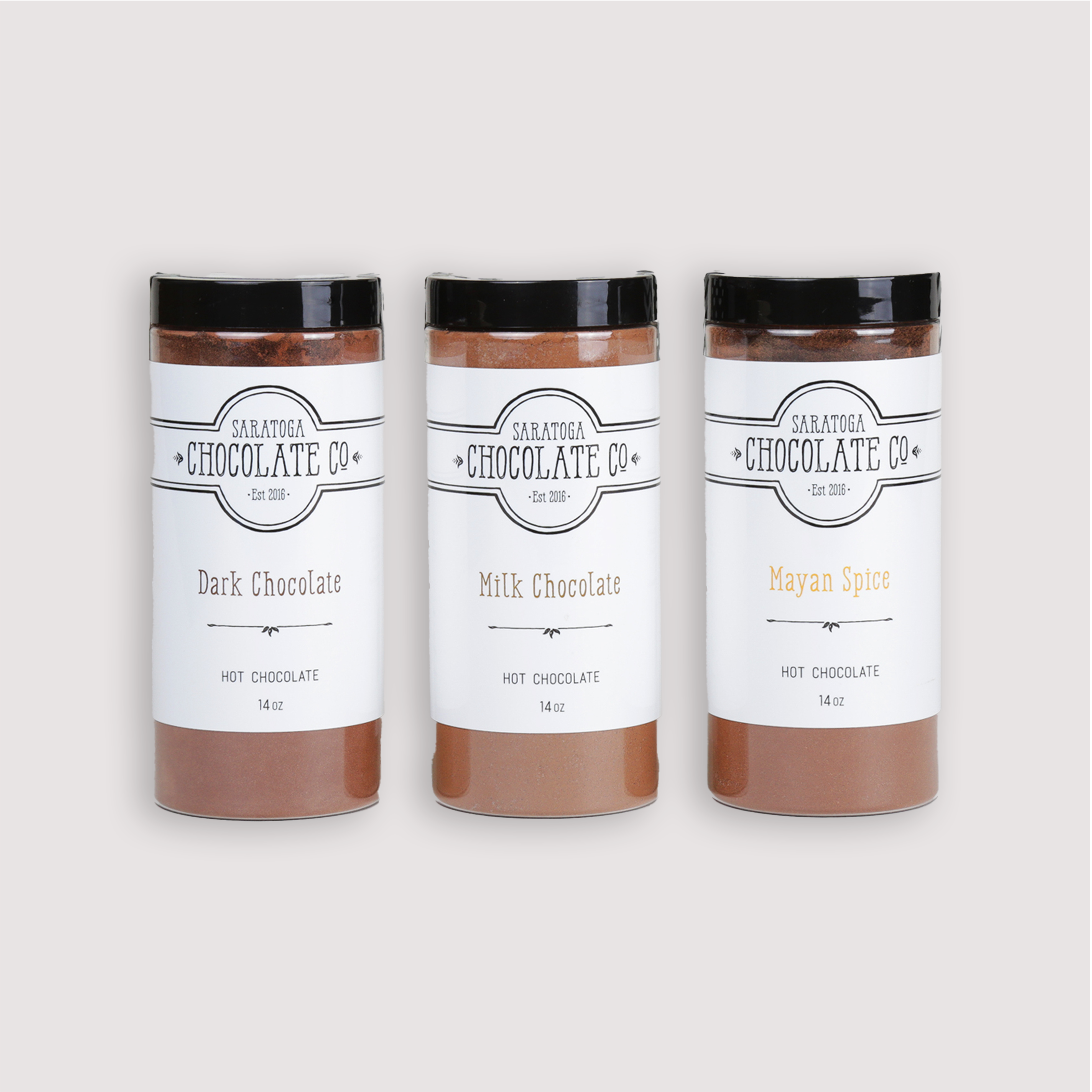 From left to right: Saratoga Chocolate Co. 14-ounce jars of Dark Hot Chocolate Mix, Milk Hot Chocolate Mix, and Mayan Spice Hot Chocolate Mix. Each jar is labeled and sealed, showcasing the variety of gourmet drinking chocolate flavors. Hot Chocolate Mix, and Mayan Spice Hot Chocolate Mix are gluten and dairy free. Milk Hot Chocolate Mix is gluten free.