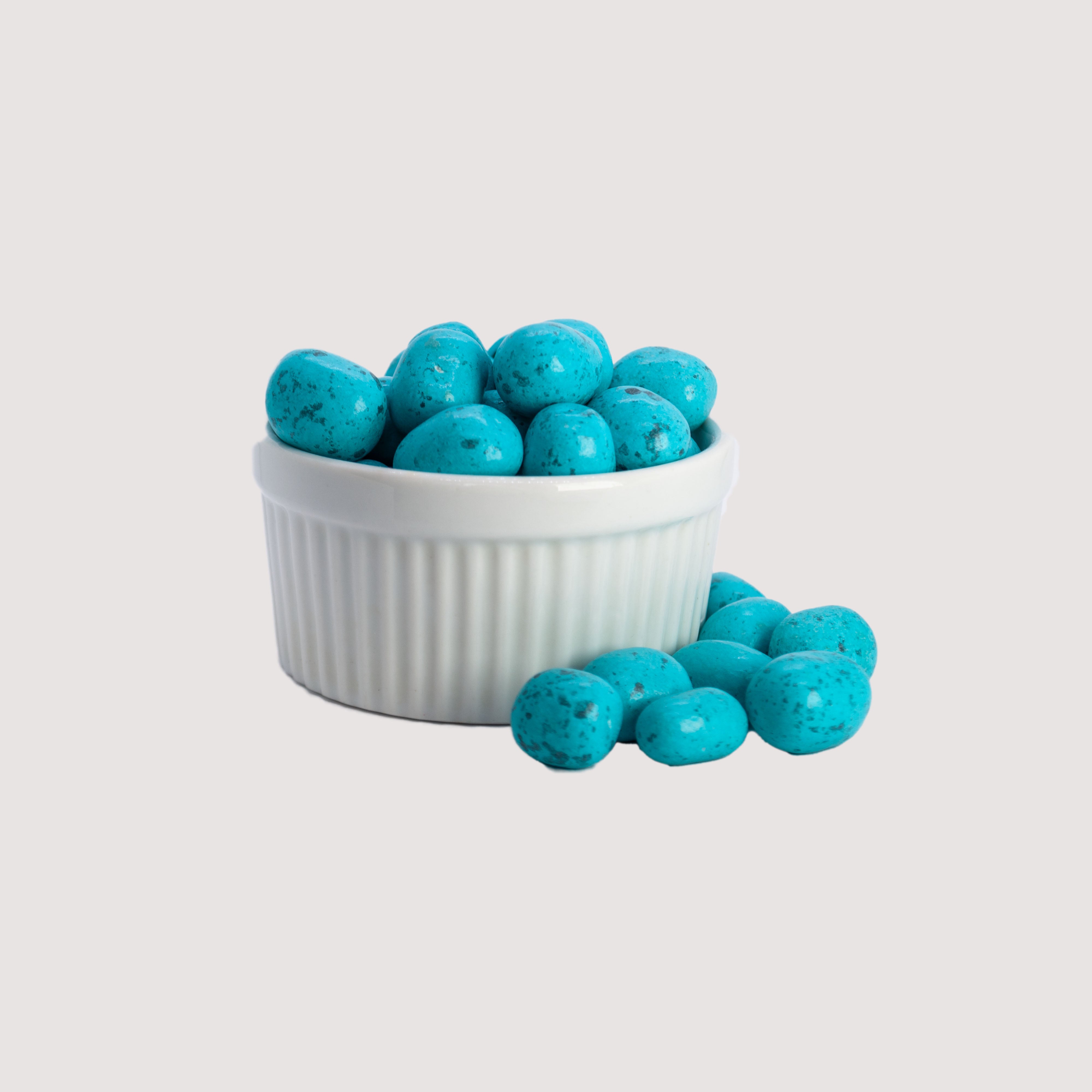 Image shows a pile of festive blue candy coated chocolate dipped mini marshmallows displayed in a fluted white bowl with some blue candy coated chocolate dipped mini marshmallows scattered in front