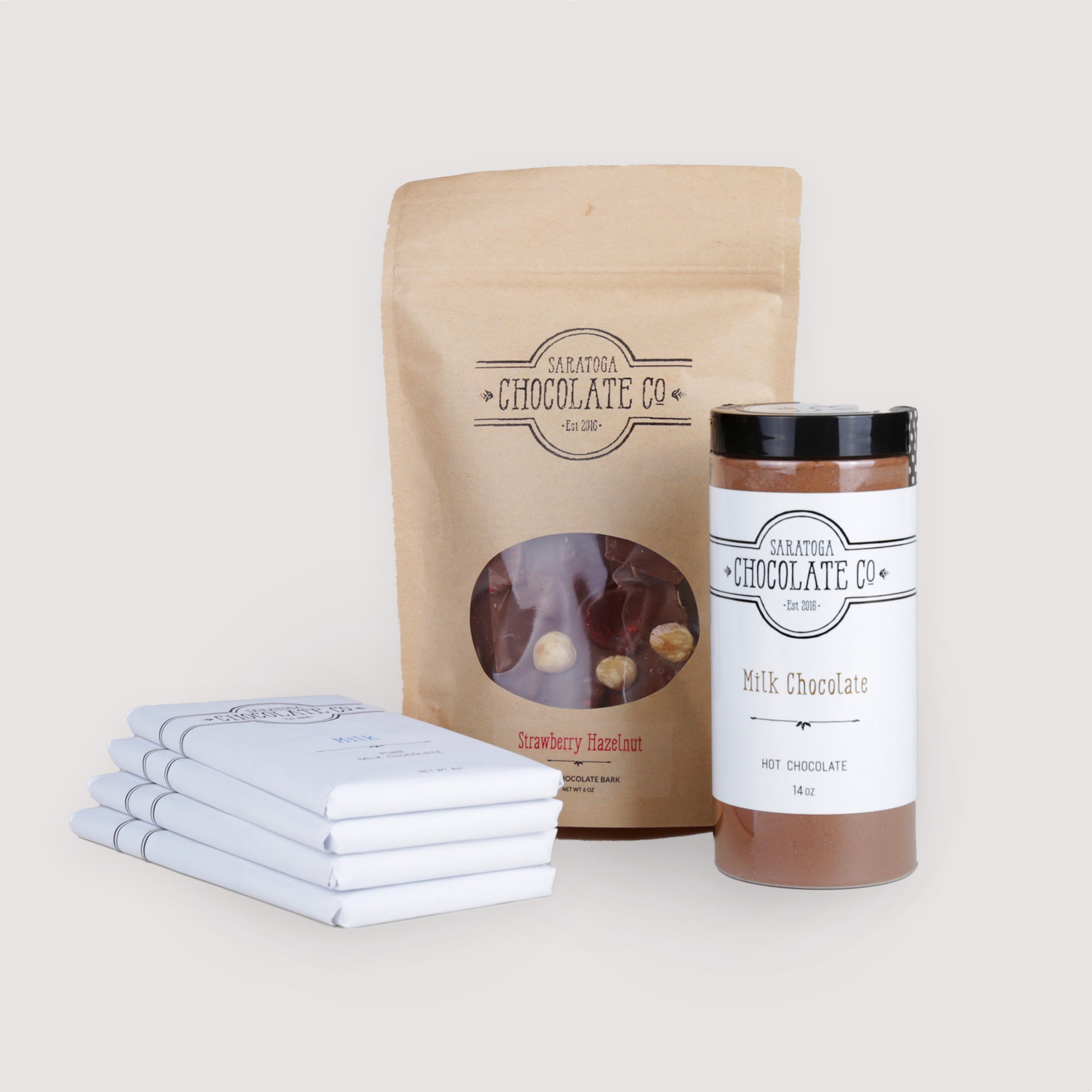 Saratoga Chocolate Co. Milk Chocolate Collection featuring four 4-ounce milk chocolate bars—Milk, Bodhi, Toasted Sesame, and Fleur de Lait—each wrapped in signature white packaging with branded labels. Includes a 6-ounce Strawberry Hazelnut Bark in a kraft paper zipper-sealed bag with a clear window displaying strawberry and hazelnut inclusions, and a 14-ounce jar of Milk Hot Chocolate mix labeled and sealed. An indulgent gluten-free assortment for milk chocolate enthusiasts.