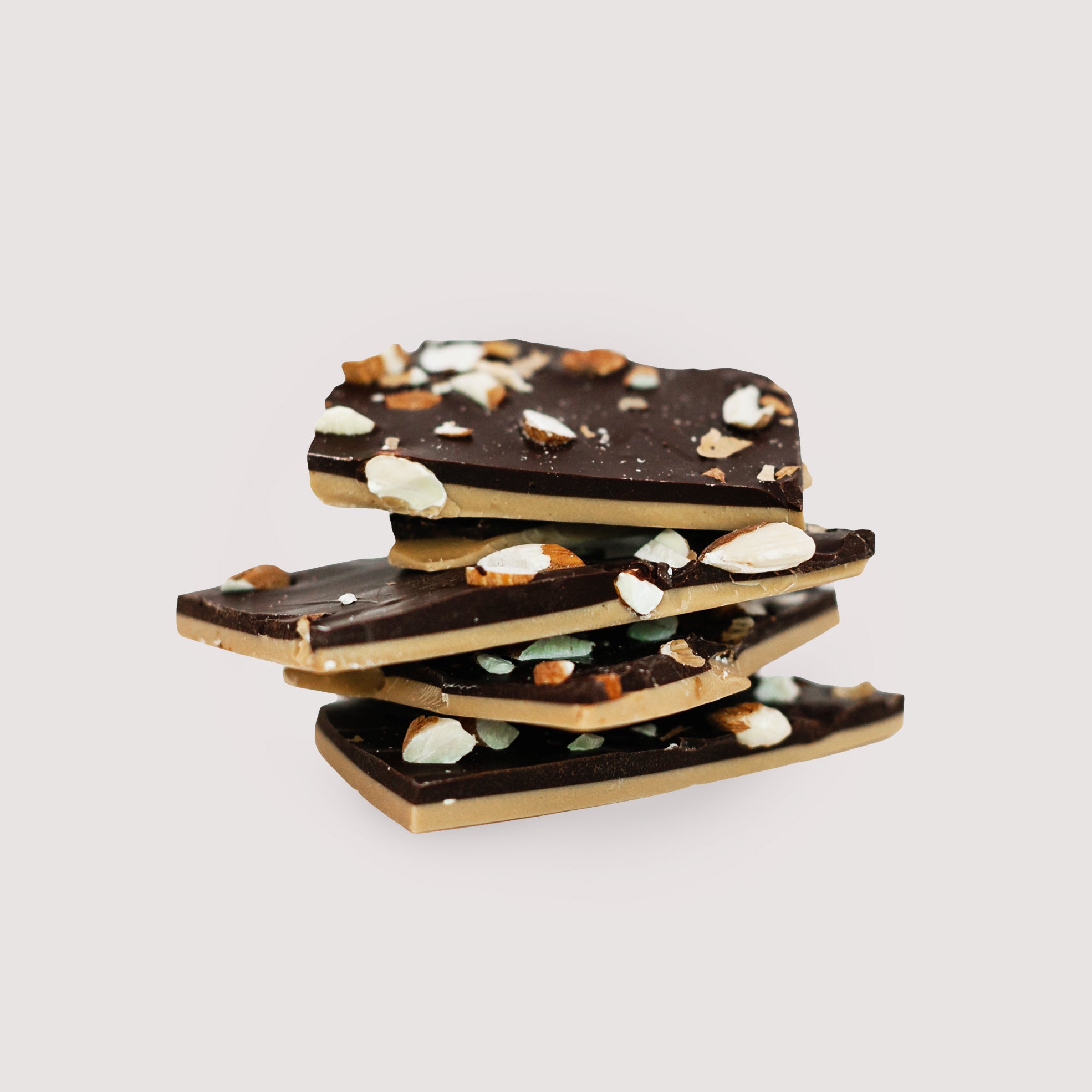 Stack of Saratoga Chocolate Co. Salted Almond Dark Chocolate Bark pieces, featuring creamy salted caramel chocolate layered with rich dark chocolate topped with roasted almonds. Saratoga Chocolate Co. Salted Almond Dark Chocolate Bark is packaged in a clear cellophane bag with a twist tie closure displaying the bark’s layers of chocolate and almond inclusions. Each bag is sealed and labeled with the Saratoga Chocolate Co. logo product name and ingredients on the back of the bag. This bark is gluten free.