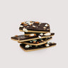 Stack of Saratoga Chocolate Co. Salted Almond Dark Chocolate Bark pieces, featuring creamy salted caramel chocolate layered with rich dark chocolate topped with roasted almonds. Saratoga Chocolate Co. Salted Almond Dark Chocolate Bark is packaged in a clear cellophane bag with a twist tie closure displaying the bark’s layers of chocolate and almond inclusions. Each bag is sealed and labeled with the Saratoga Chocolate Co. logo product name and ingredients on the back of the bag. This bark is gluten free.