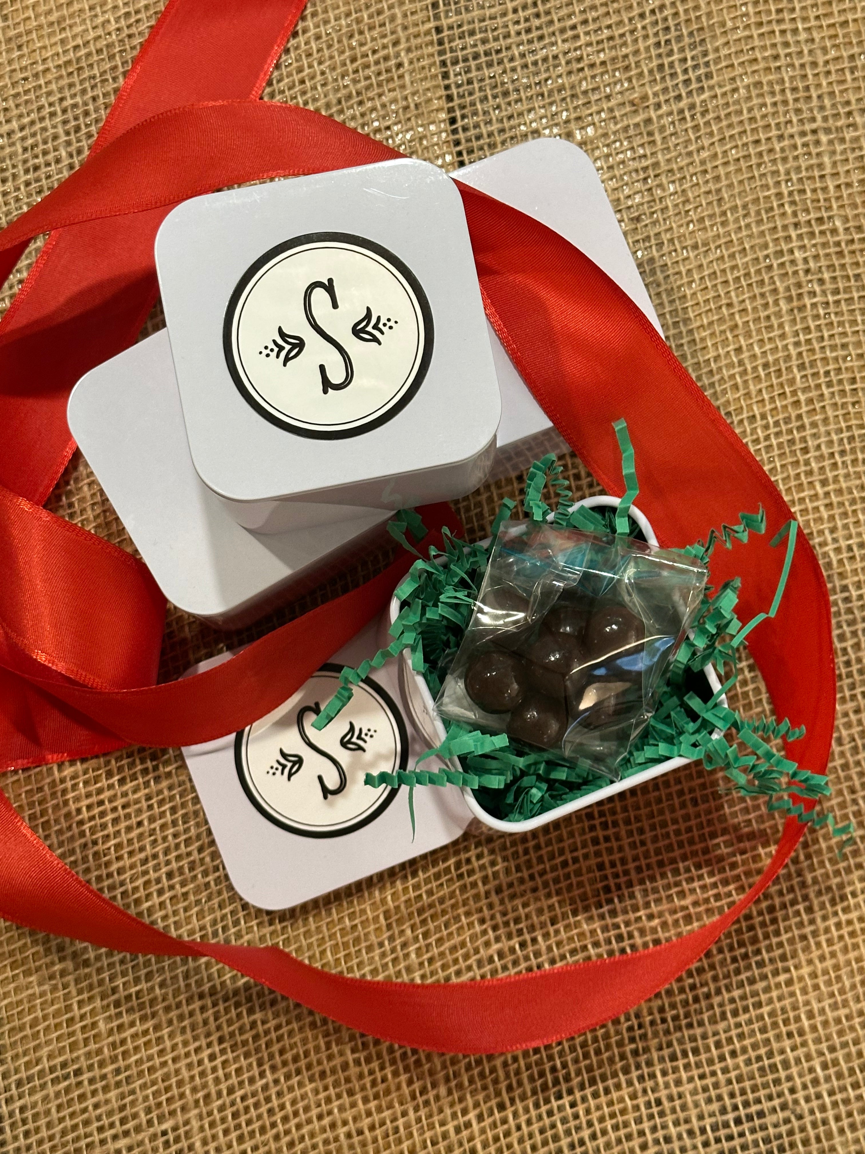 Close-up of small white square tins on a burlap surface. One tin is open, showing green crinkle filler and several round dark chocolates in a cellophane bag. A red ribbon is draped across the tins