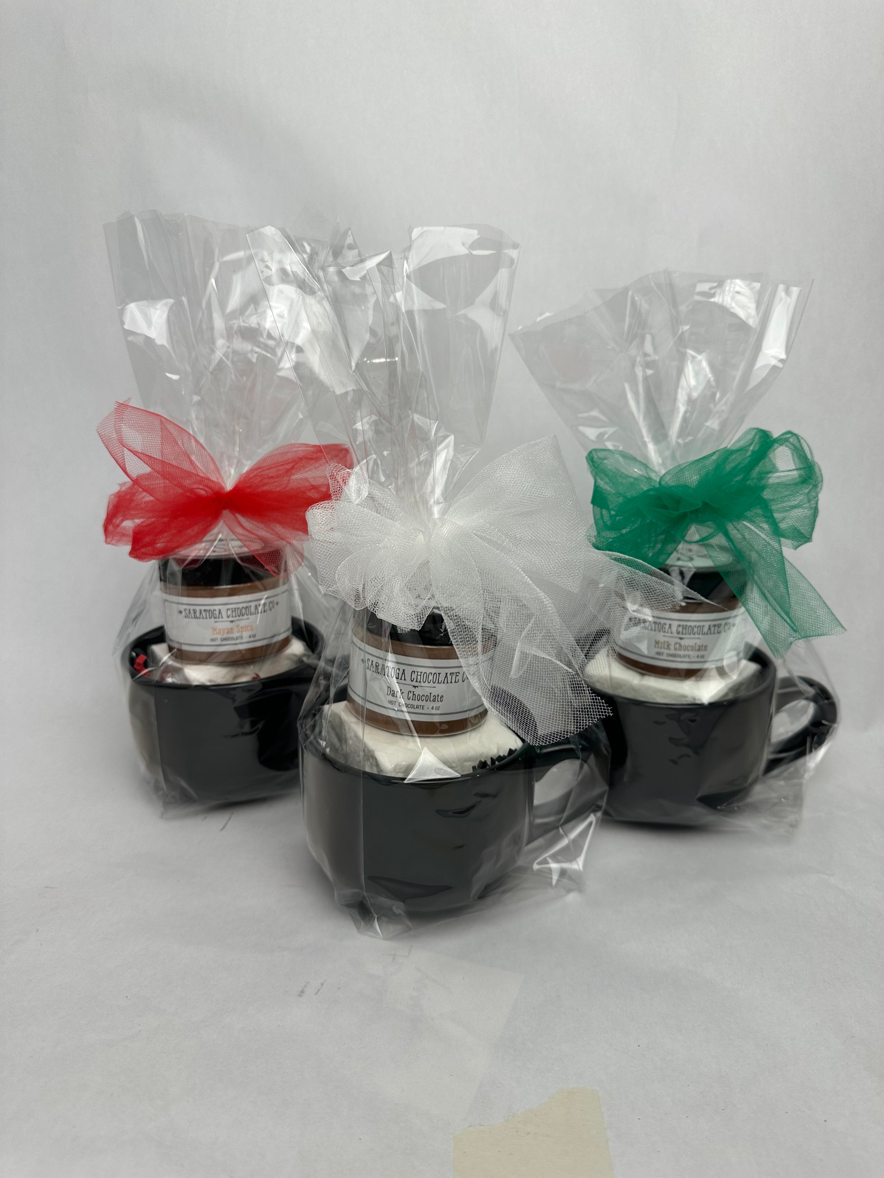 Three cozy hot cocoa gift bundles in matte black mugs, each wrapped in clear cellophane and tied with tulle bows — red, white, and green — showing a jar of drinking chocolate and a jumbo marshmallow inside. 


