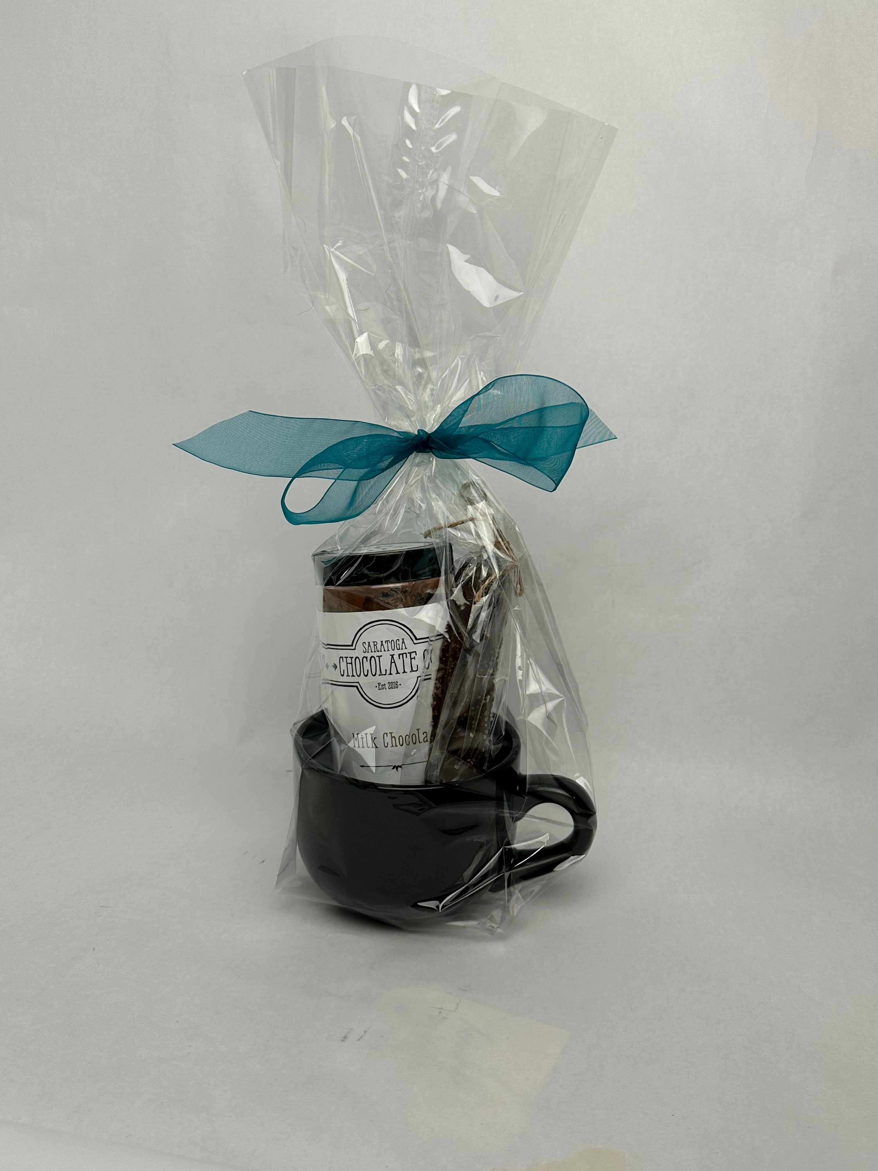 Glossy black stoneware mug packaged as a gift, wrapped in clear cellophane with a teal bow. Inside is a full-size jar of gourmet milk chocolate drinking cocoa and a dark chocolate caramel stir stick coated in sea salt.