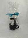 Glossy black stoneware mug packaged as a gift, wrapped in clear cellophane with a teal bow. Inside is a full-size jar of gourmet milk chocolate drinking cocoa and a dark chocolate caramel stir stick coated in sea salt.