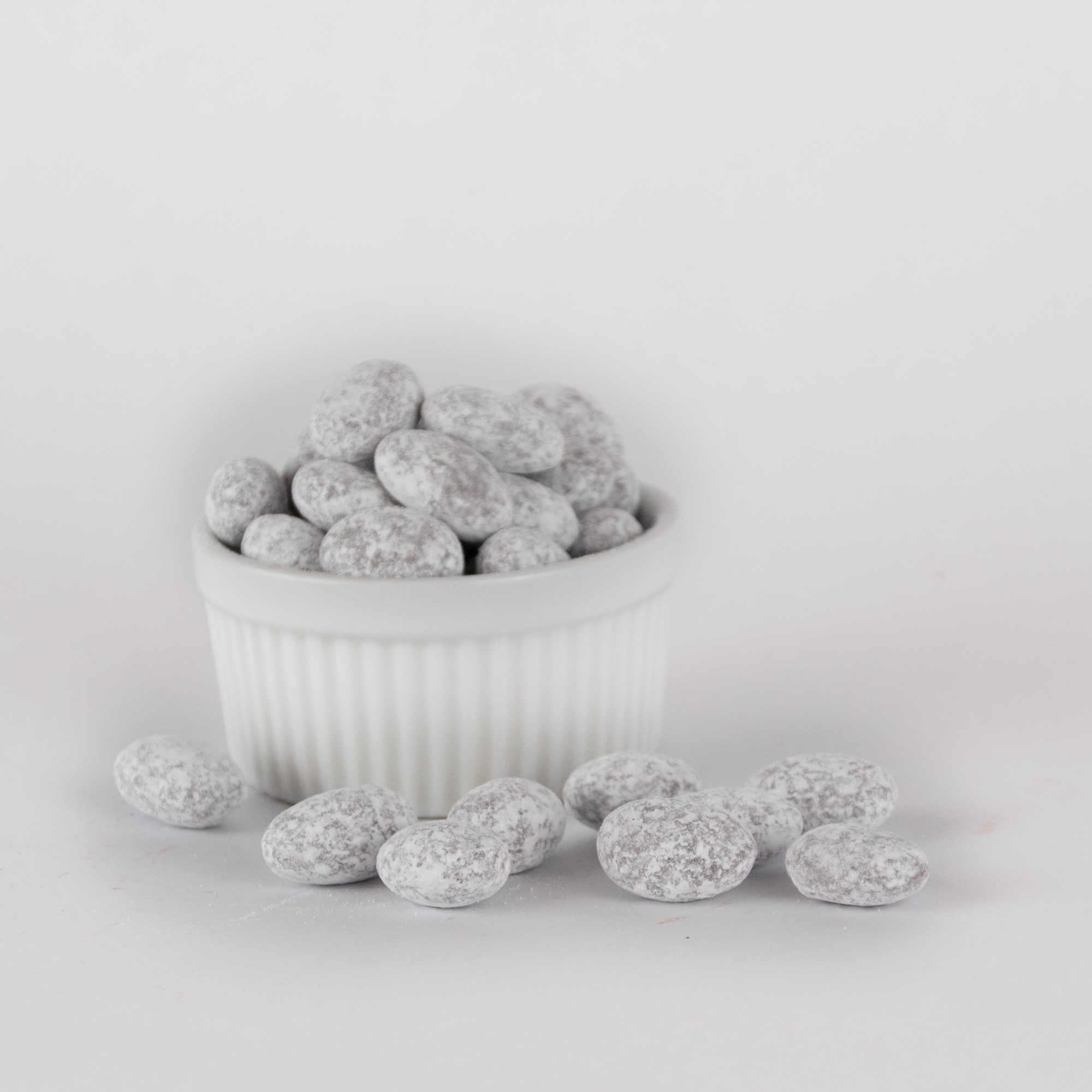 Powdered toffee-coated almonds displayed in a fluted white bowl, with several pieces scattered in front, showcasing their light, powdery coating and irregular texture.
