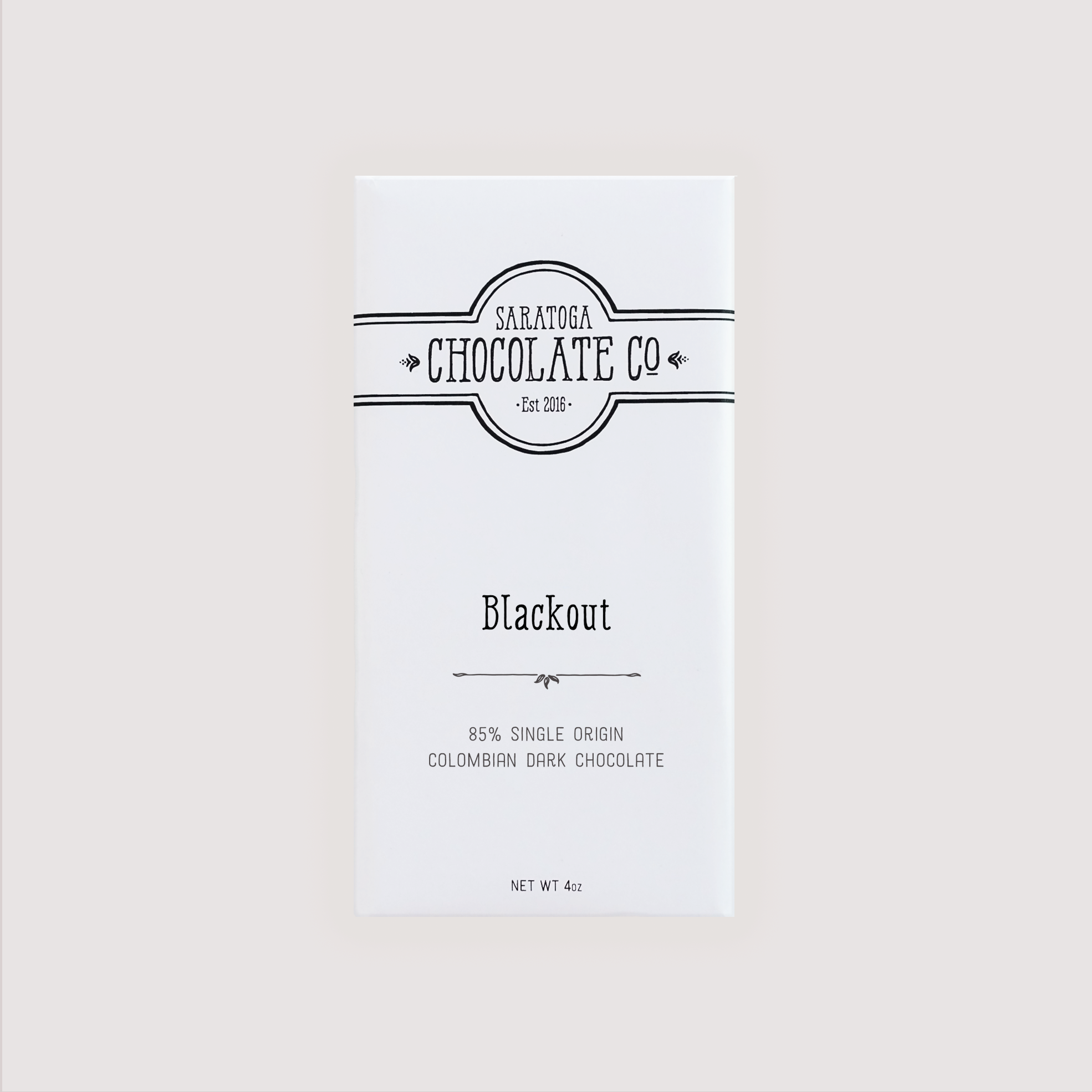 Front of Saratoga Chocolate Co. Blackout Bar wrapped in a white paper package with a black flavor label. Label features the Saratoga Chocolate Co. logo and describes the bar as 85% single origin Colombian dark chocolate from the Tumaco River Valley. This bar is gluten and dairy free.