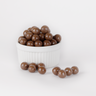 Image shows a pile of small, round spheres of milk chocolate coated sea salt caramels displayed in a fluted white bowl with some milk chocolate coated sea salt caramels scattered in front.