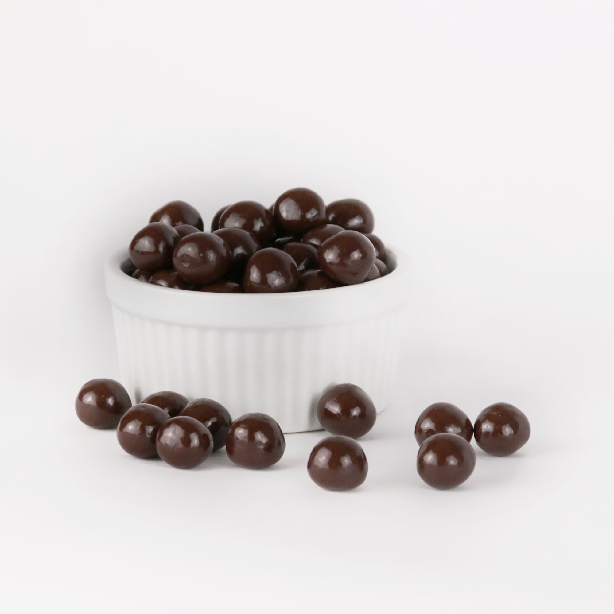 Image shows a pile of small, round spheres of dark chocolate coated sea salt caramels displayed in a fluted white bowl with some dark chocolate coated sea salt caramels scattered in front.