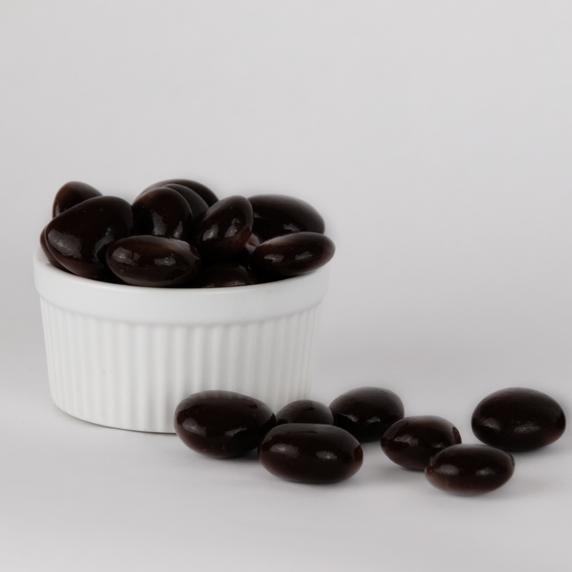 Image shows a pile of dark chocolate brandied cherries displayed in a fluted white bowl with some chocolate covered brandied cherries scattered in front of the bowl.