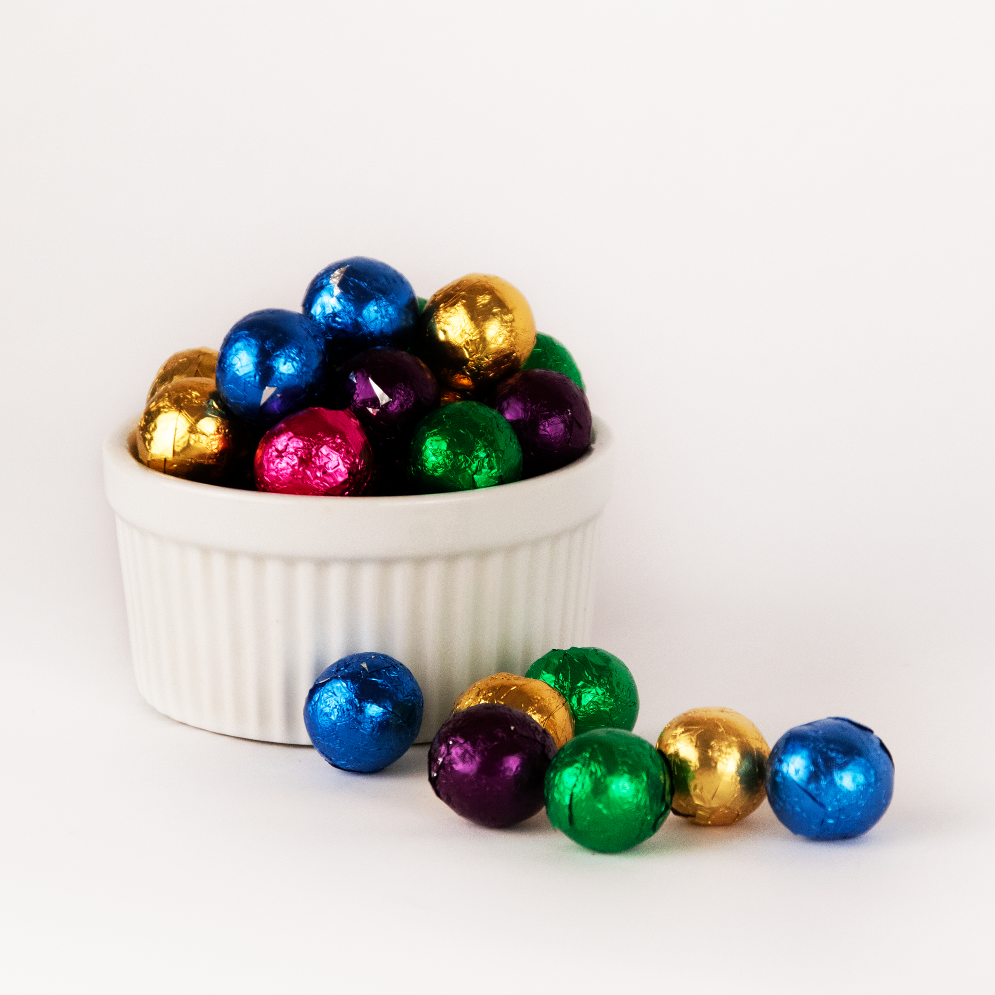 Image shows a pile of small, round spheres of dark chocolate balls wrapped in brightly colored gold, blue, green, pink and purple foil wrappers displayed in a fluted white bowl with pile of small, round spheres of dark chocolate balls wrapped in brightly colored foils scattered in front.