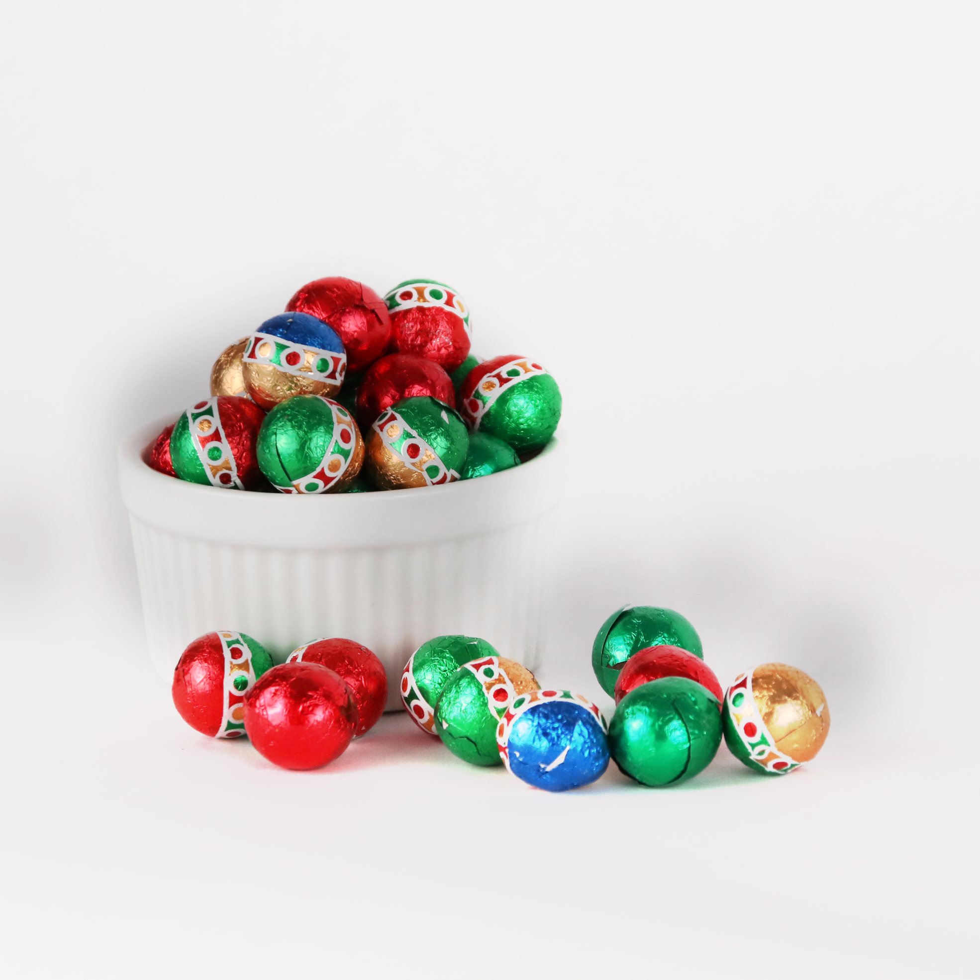 Milk chocolate foil balls wrapped in shiny red, green, and gold foil with holiday ornament, displayed in a fluted white bowl with several scattered in front, reflecting light with a festive appearance.