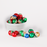 Milk chocolate foil balls wrapped in shiny red, green, and gold foil with holiday ornament, displayed in a fluted white bowl with several scattered in front, reflecting light with a festive appearance.