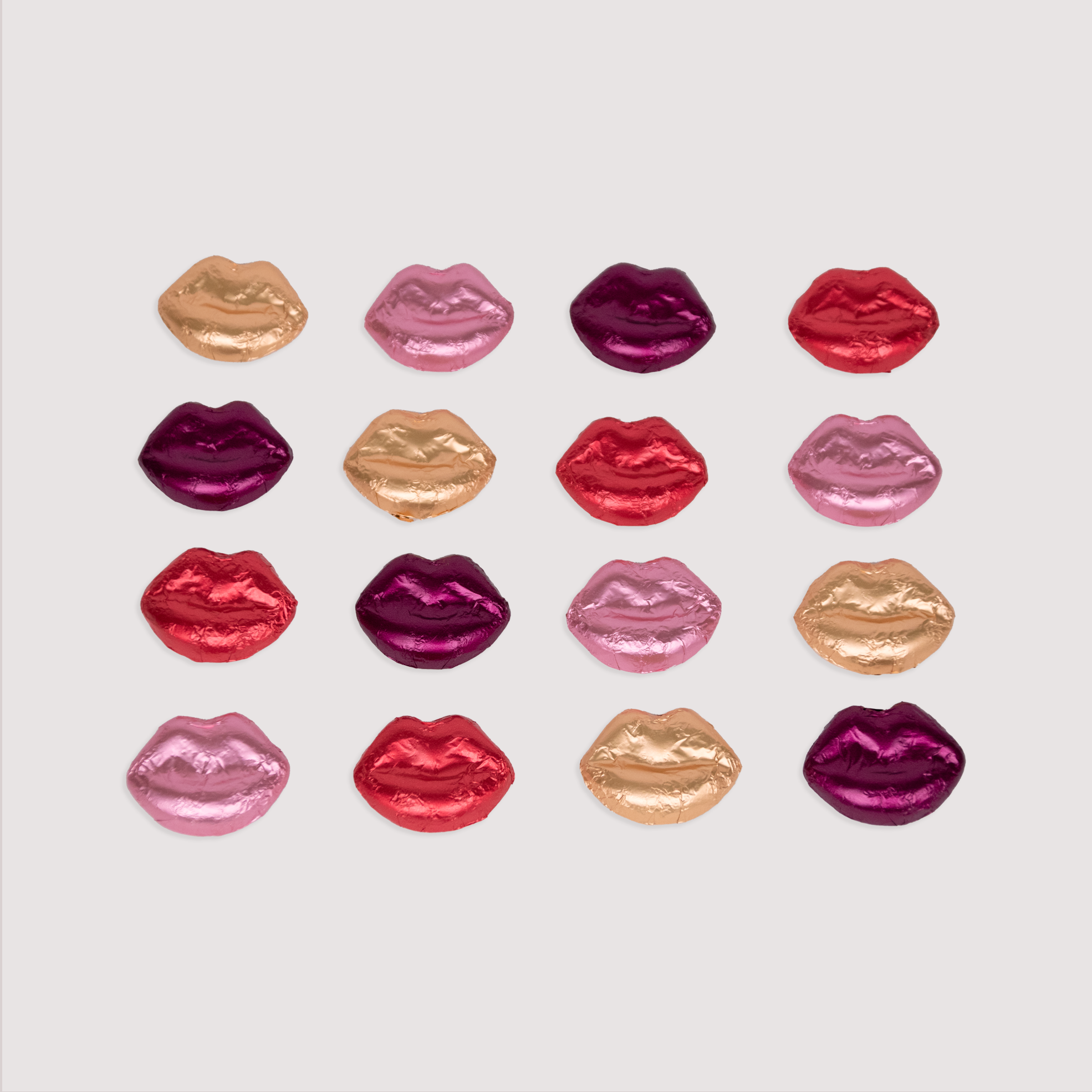 Foil-wrapped chocolate lips by Saratoga Chocolate in vibrant red, pink, and purple colors, scattered playfully on a light pink background.