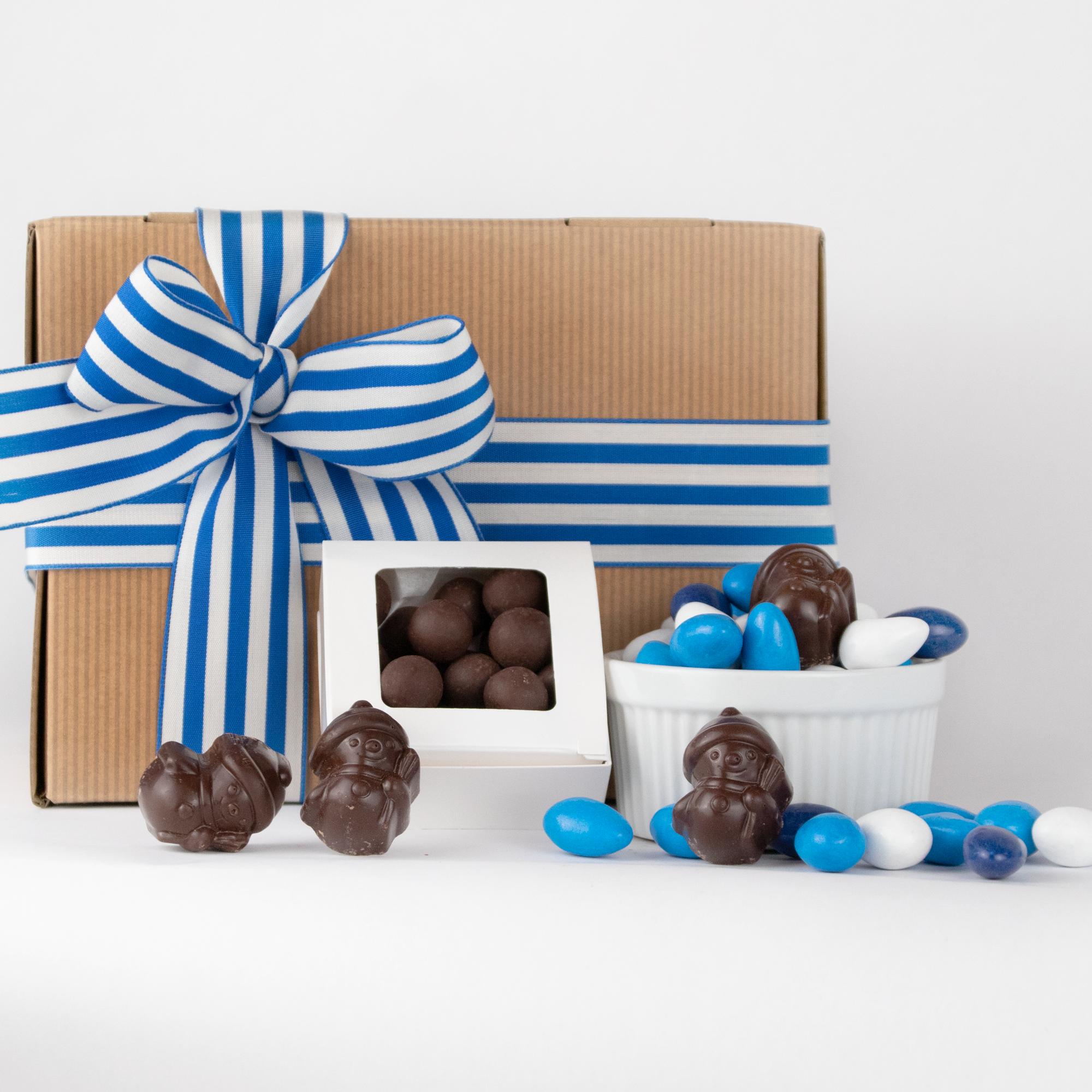 Hanukkah gift set by Saratoga Chocolate Co featuring almonds coated in white, light blue, and dark blue candy shells, four chocolate snowmen, a white box with a clear lid filled with assorted chocolate balls, and a light brown gift box tied with a blue and white ribbon, all arranged with festive Hanukkah-themed accents.