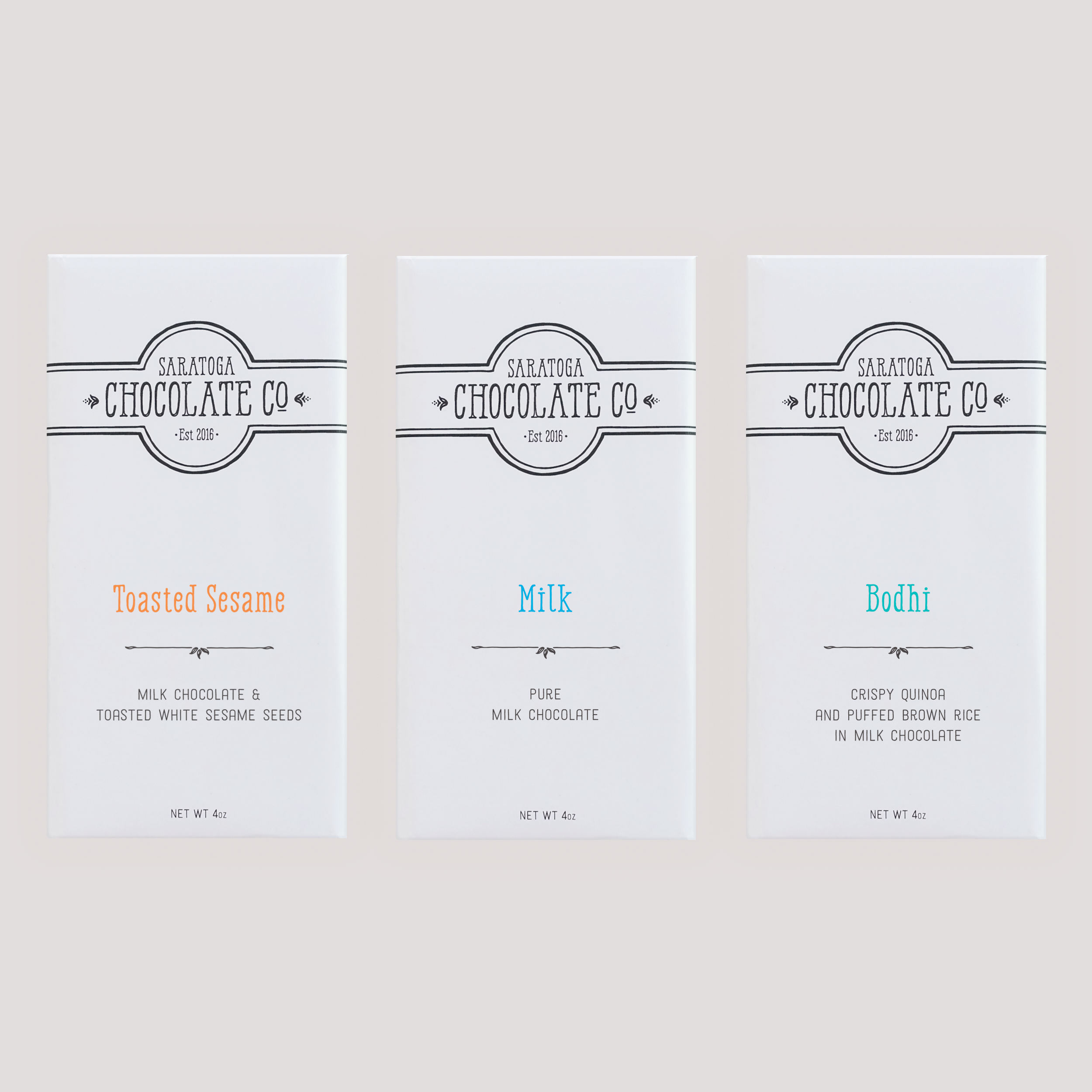 Three Saratoga Chocolate Co. milk chocolate bars in white wrappers with distinct labels: Toasted Sesame, Milk and Bodhi; arranged side by side on a light background.