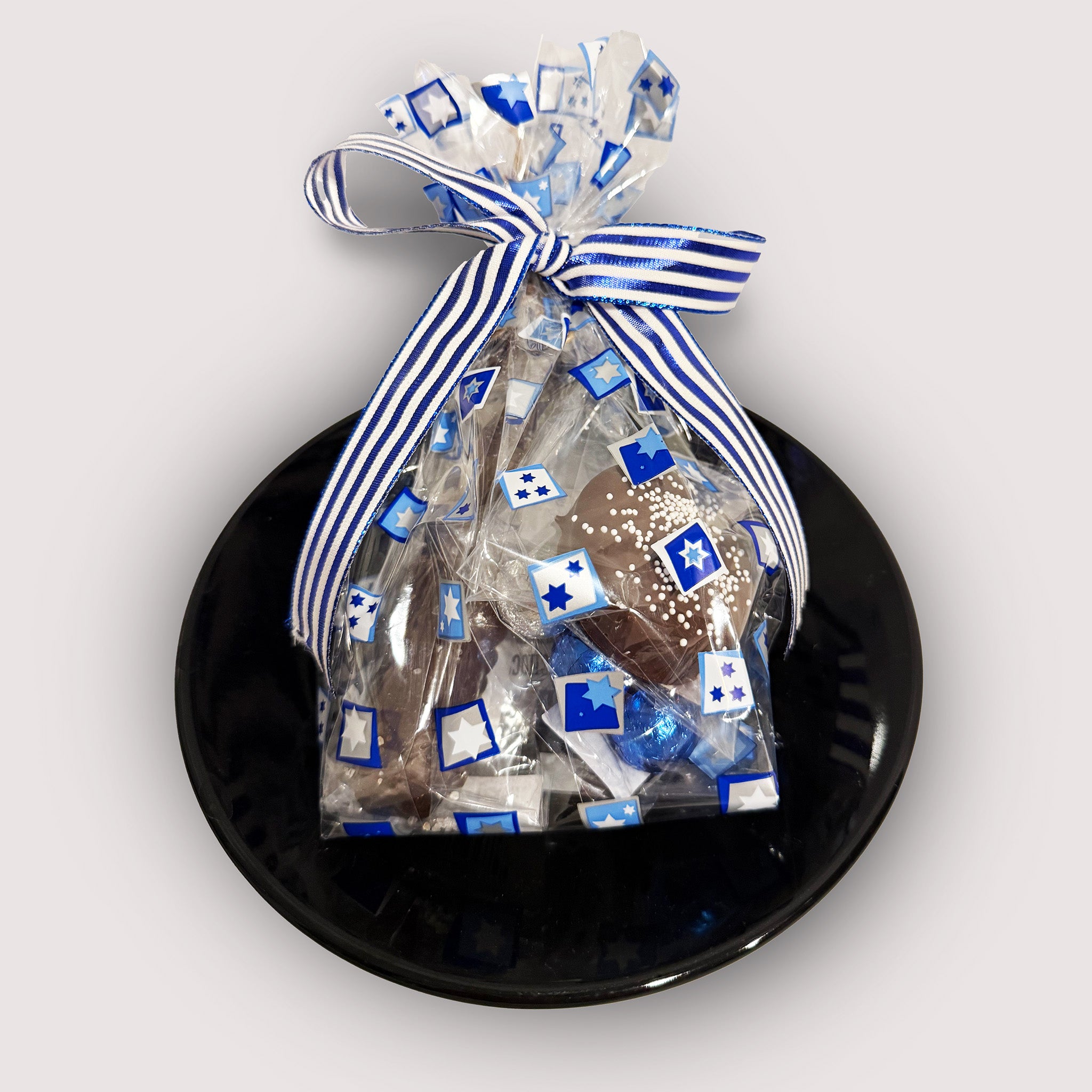 A clear bag with chocolate treats with blue stars and a striped blue ribbon on top. Displayed on a black plate and neutral background.