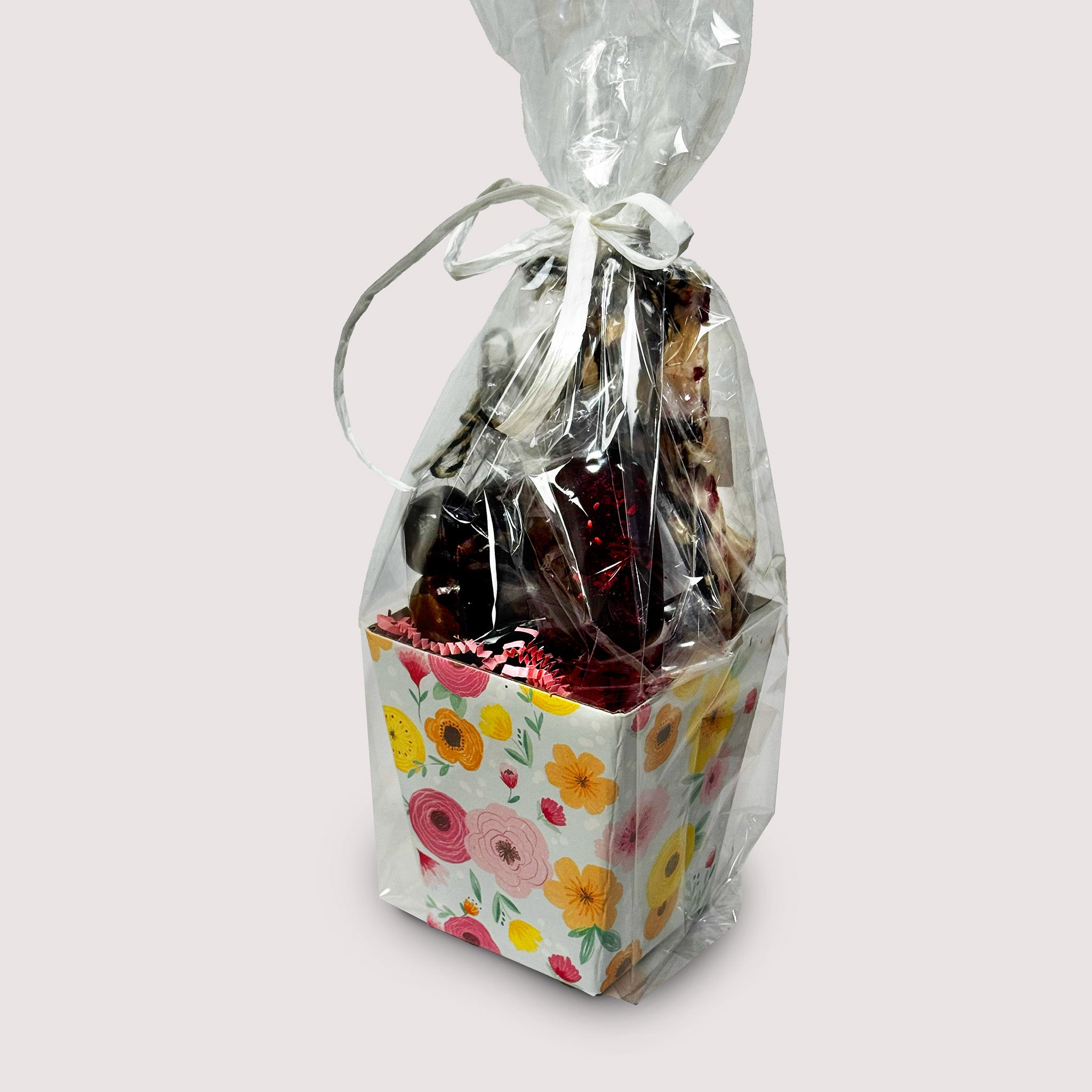 A gift bundle in floral basket and chocolate treats in a clear bag with white ribbon on a neutral background