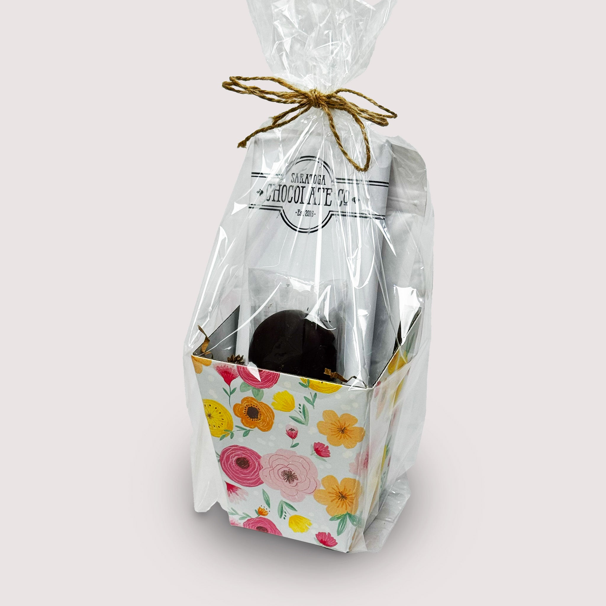 Floral box from Saratoga Chocolate Co., featuring a Latte Bar, Chocolate-Covered Espresso Beans, and chocolate covered Oreo in  a clear bag with twine bow tie displayed on a neutral background.