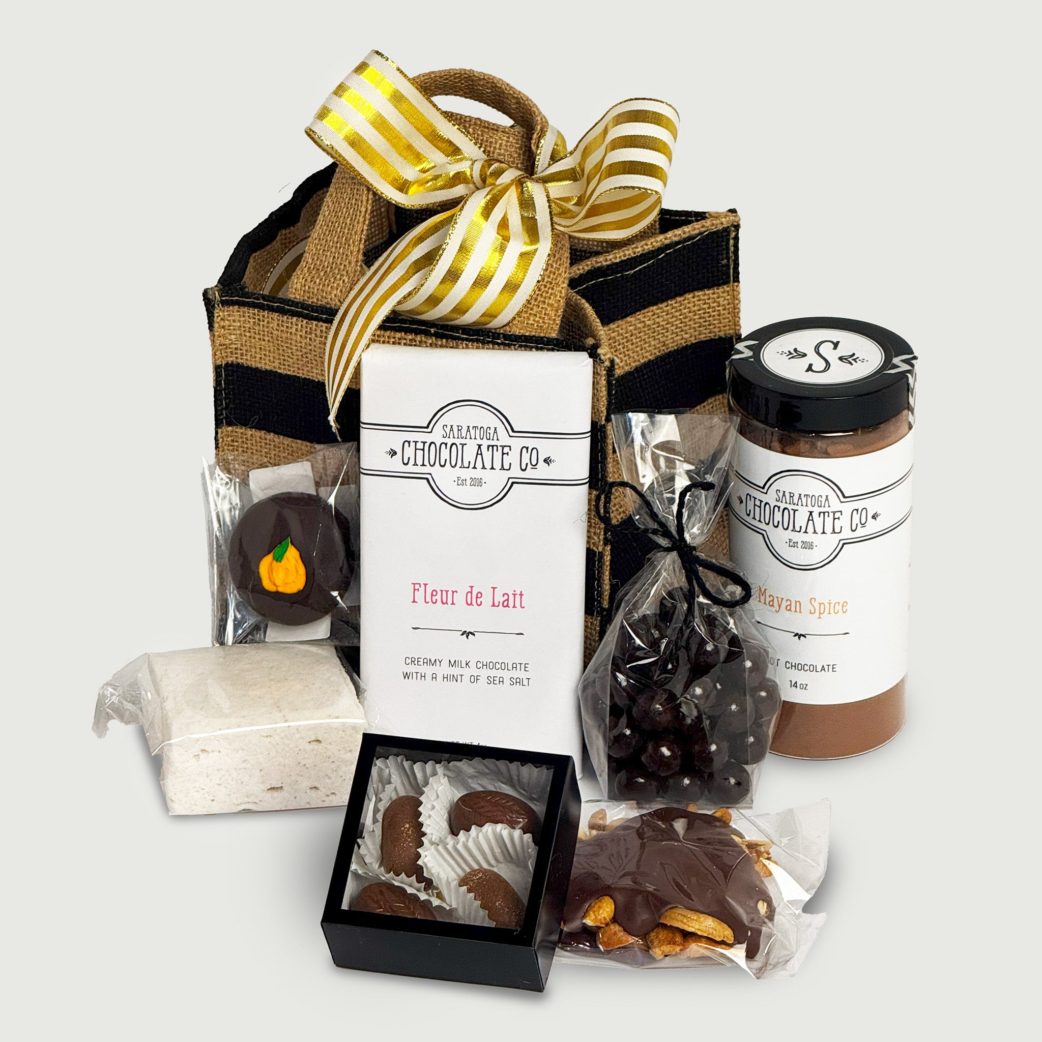 Assorted Saratoga Chocolate Co. gift box displayed on a neutral background with a tote bag tied in a golden ribbon. Includes Fleur de Lait Bar, Milk Chocolate Cashew Turtle, Pumpkin Spice Jumbo Marshmallow, Dark Chocolate Sea Salt Caramels, Mayan Spice Hot Chocolate, dark chocolate dipped Oreos, and a 4-pack truffle box with apple cider and pumpkin spice caramel truffles.