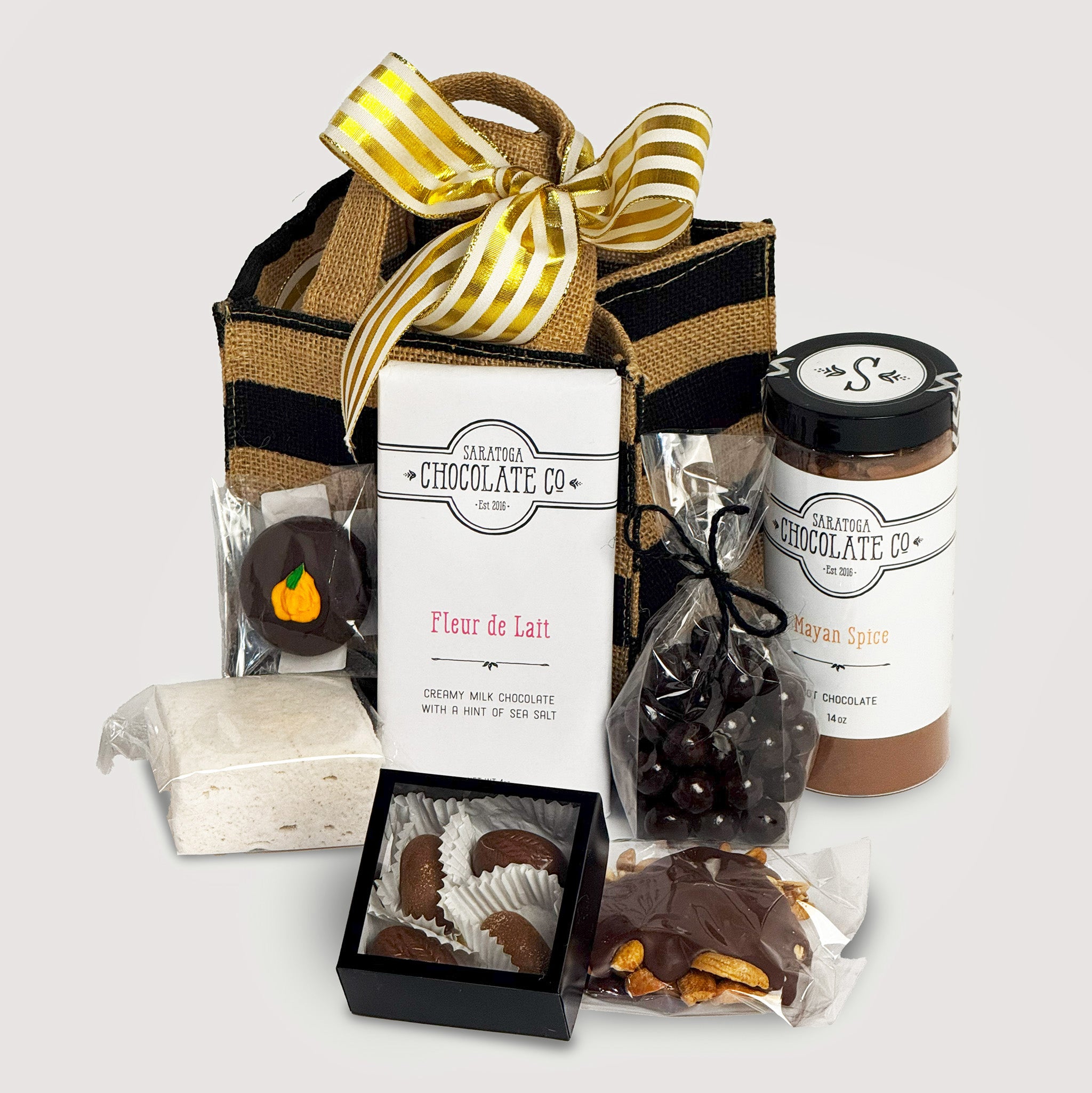 Chocolate gift set with a striped bow, including Fleur de Lait Bar, Milk Chocolate Cashew Turtle, Pumpkin Spice Jumbo Marshmallow, Dark Chocolate Sea Salt Caramels, Mayan Spice Hot Chocolate (14 oz), Oreos dipped in dark chocolate (Velvet by SCCo), and a 4-piece truffle box featuring Apple Cider and Pumpkin Spice Caramel truffles. Beautifully presented in front of a tote bag tied with a golden ribbon, on a light gray background.