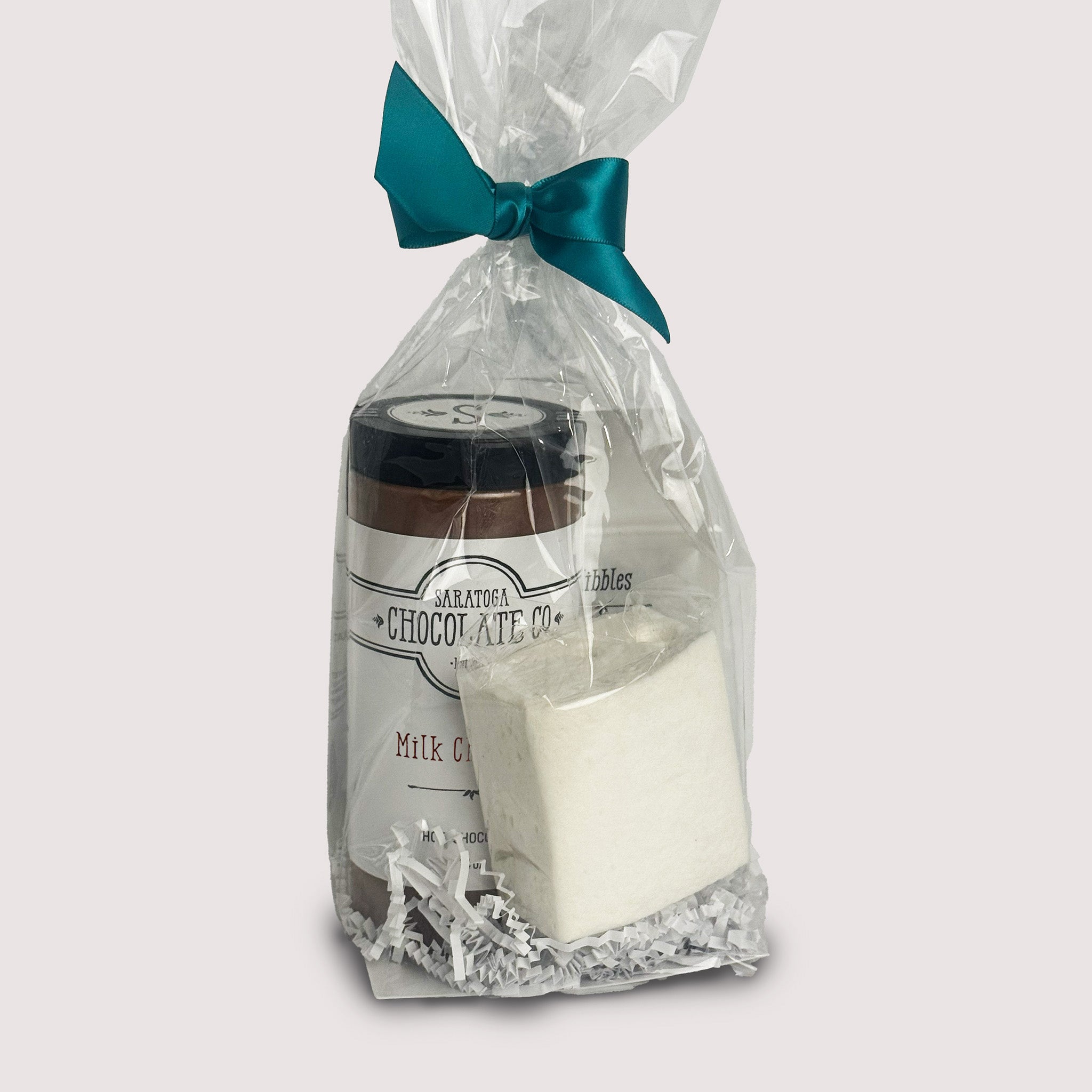 A jar of hot chocolate, small Nibble pouch and a white marshmallow in clear packaging with a teal ribbon on a light gray background