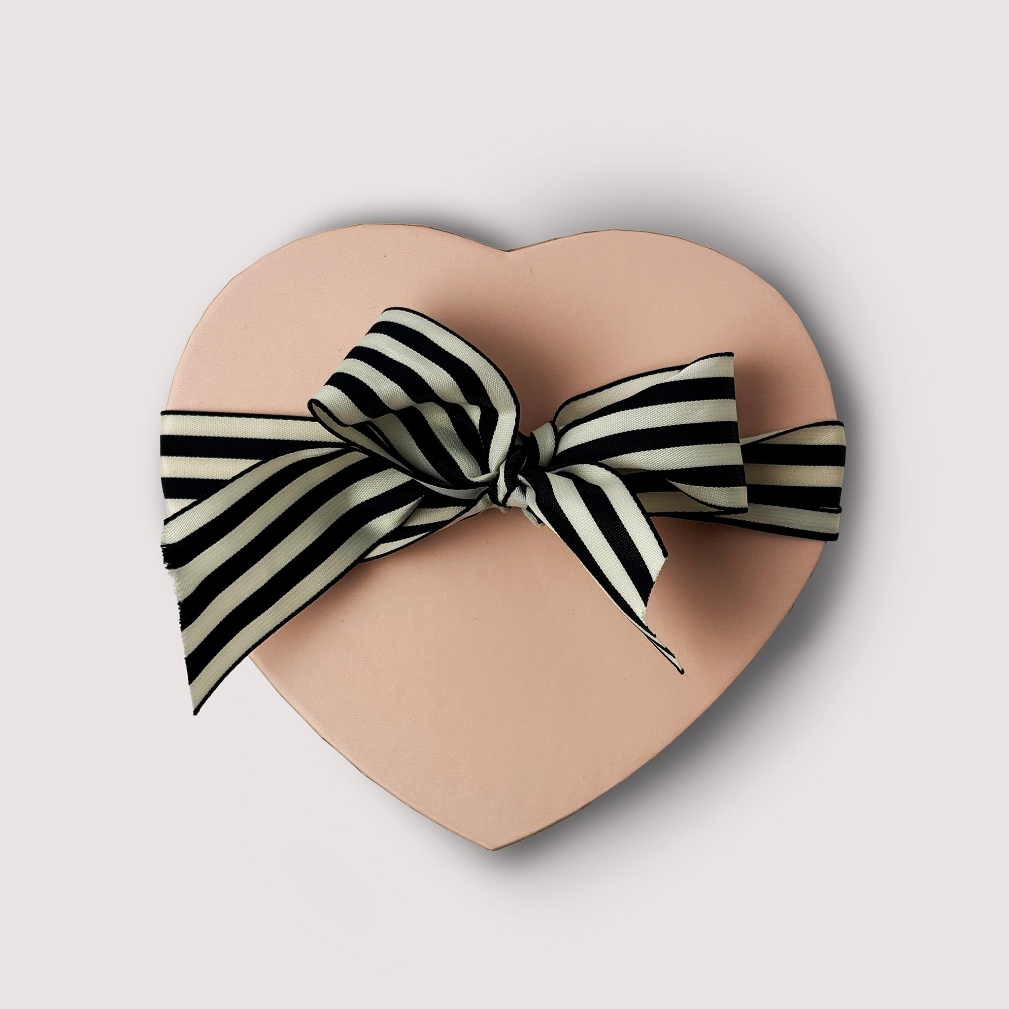 A Pink heart shape box of chocolate treats with black and white striped ribbon tide in a bow displayed on a neutral background