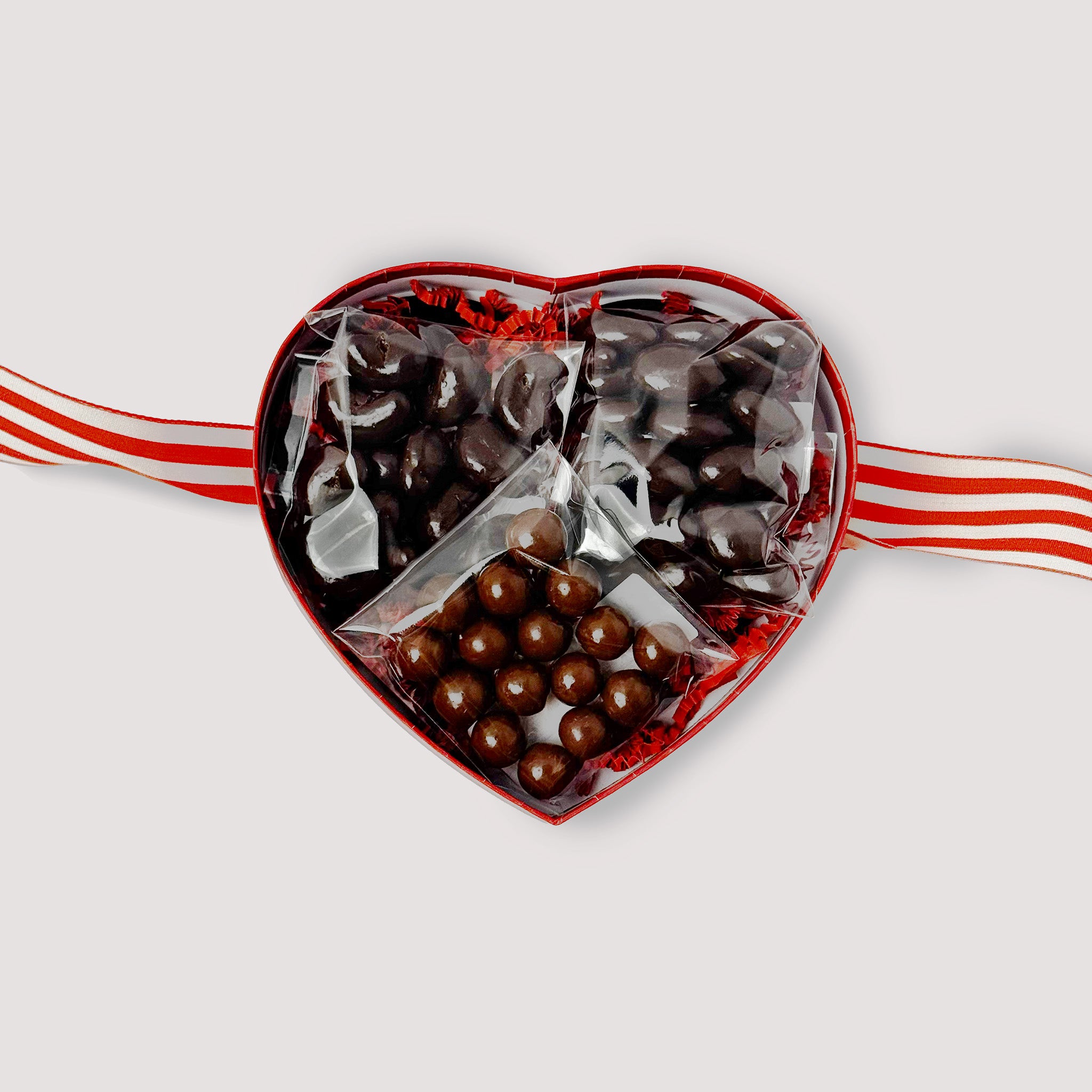 Heart-shaped chocolate gift set with chocolate covered cashews, almonds and caramels with a ribbon on a light gray background