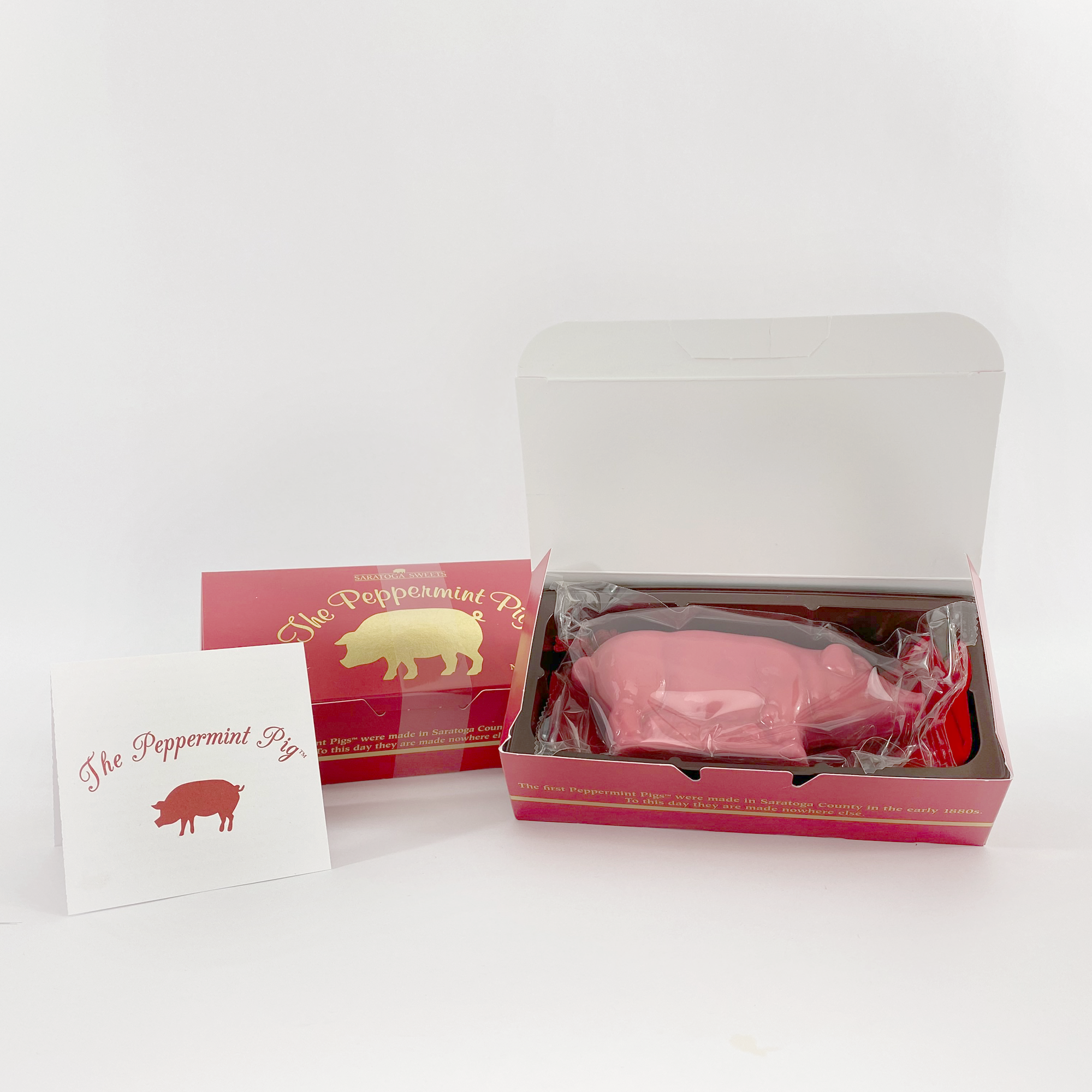 A red box containing a pink peppermint pig candy wrapped in clear plastic, accompanied by a white 'The Peppermint Pig' postcard, all part of a festive holiday tradition.