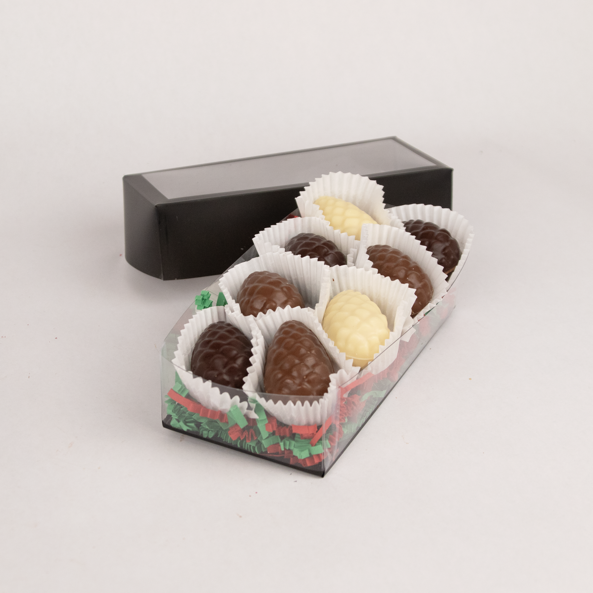 Clear gift box of eight Saratoga Chocolate Co. milk chocolate, dark chocolate and white chocolate pinecone-shaped praline bonbons, each nestled in white candy cups on colorful paper crinkle grass. The box features a clear lid with an elegant black border, showcasing the artisanal chocolate presentation. This product contains nuts.

