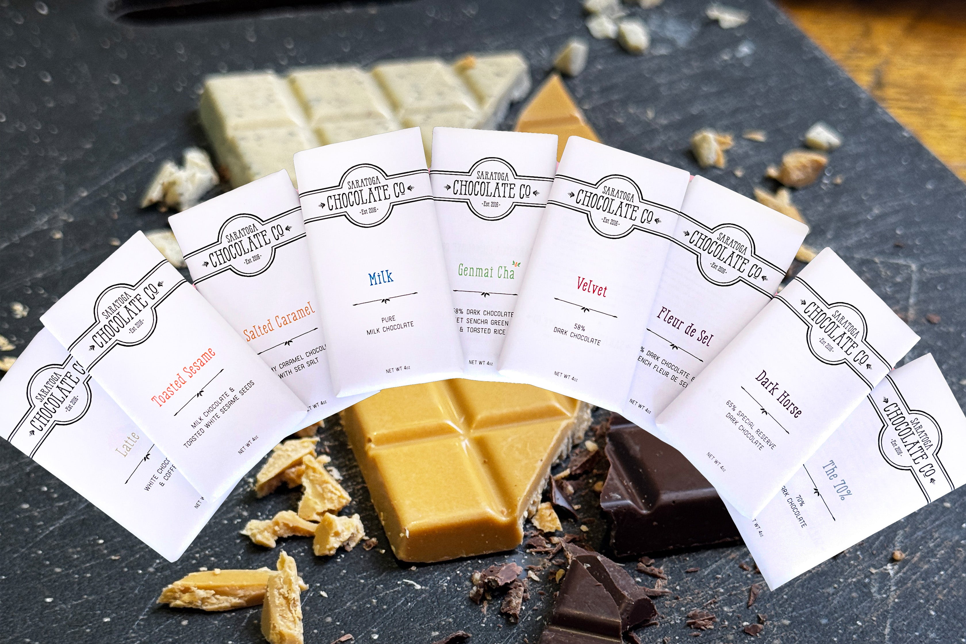 Saratoga Chocolate Co. milk and dark chocolate bars wrapped in white logo wrappers, arranged in a neat arch on a background with pieces of cut white, caramel and dark chocolate bars on a black cutting board
