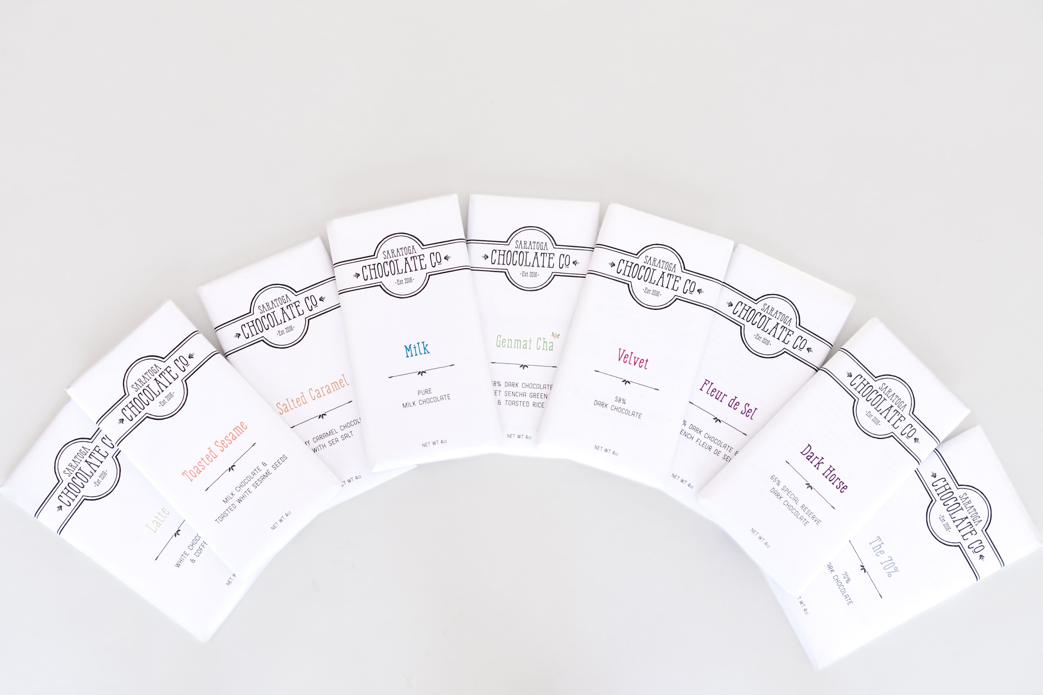 White-wrapped chocolate bars arranged in a rainbow arch, each with a dark logo of Saratoga Chocolate Co  and colorful text indicating the chocolate’s name, creating a vibrant and visually appealing display.