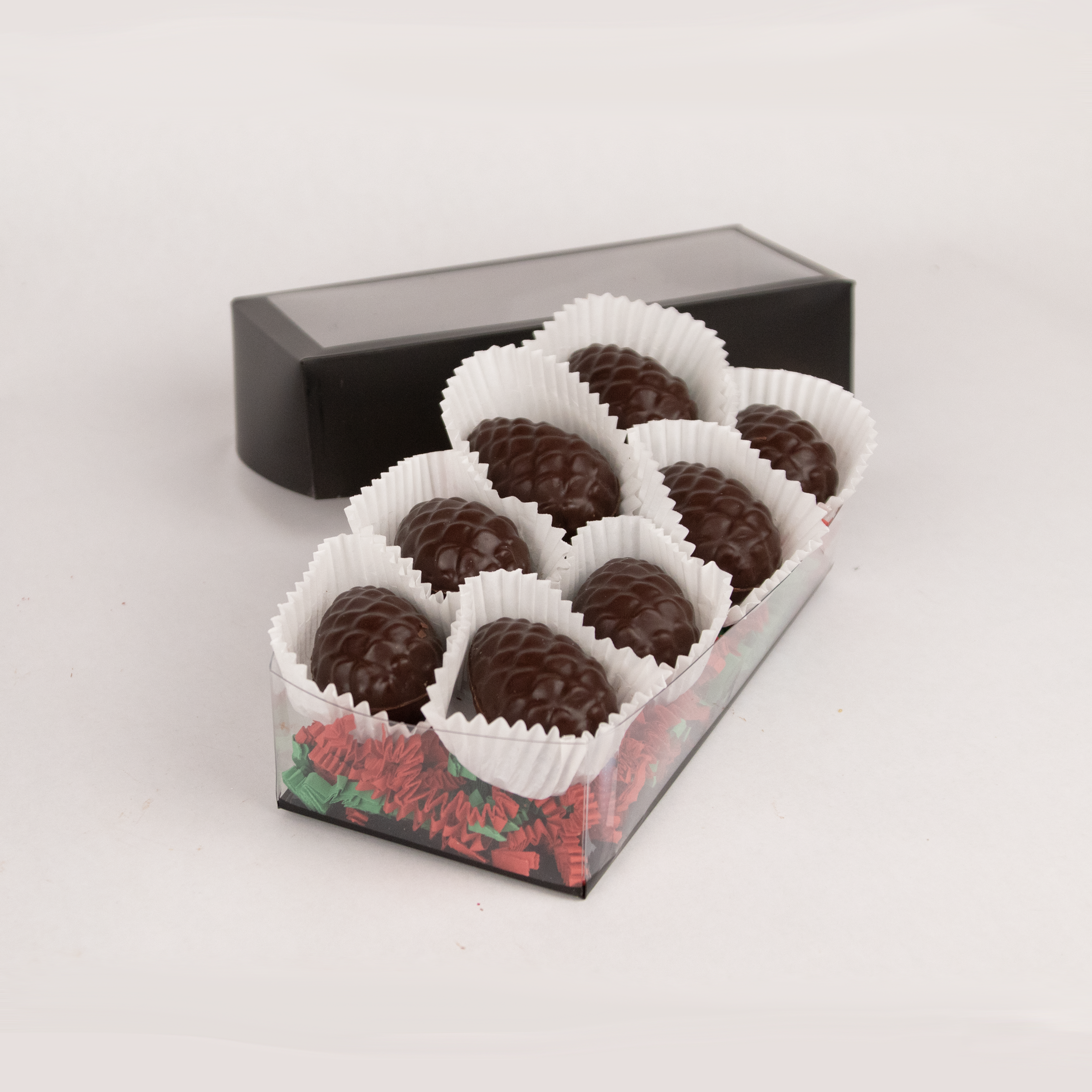 Clear gift box of eight Saratoga Chocolate Co. dark chocolate pinecone-shaped praline bonbons, each nestled in white candy cups on colorful paper crinkle grass. The box features a clear lid with an elegant black border, showcasing the artisanal chocolate presentation. This product contains nuts.