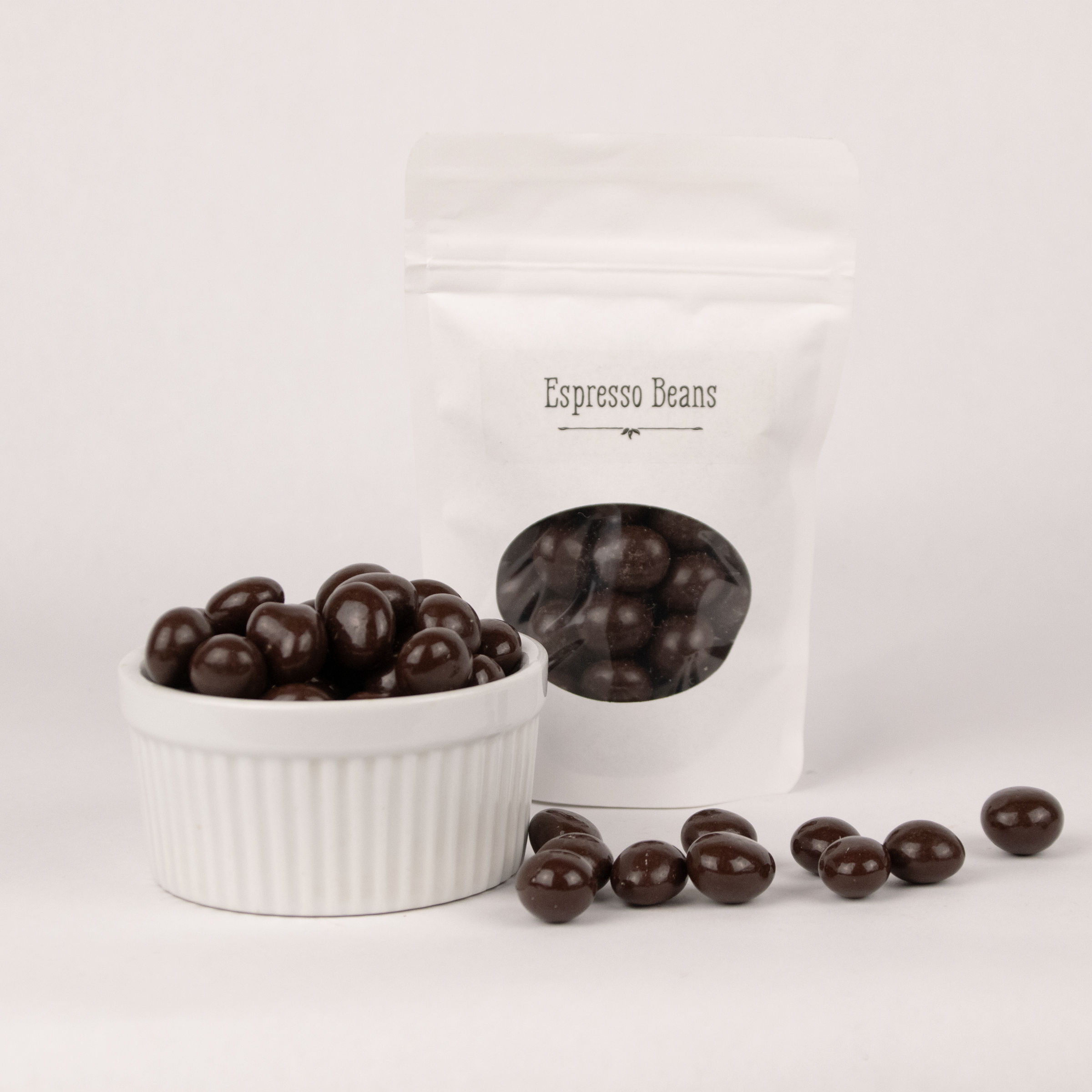 Image shows a pile of dark chocolate coated espresso beans displayed in a fluted white bowl with pile of dark chocolate espresso beans scattered in front. Behind the bowl and offset to the right is a pouch of dark chocolate covered espresso beans representing how the product is packaged.