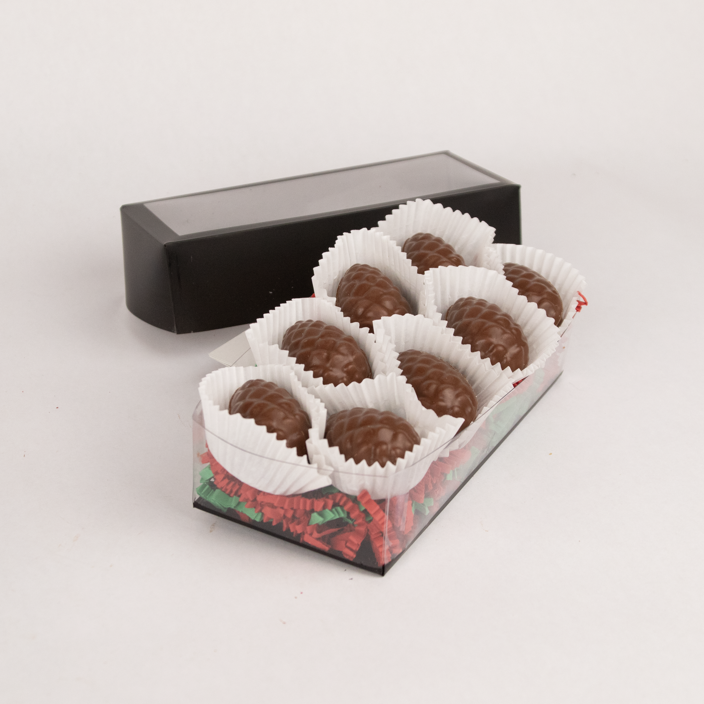 Clear gift box of eight Saratoga Chocolate Co. milk chocolate pinecone-shaped praline bonbons, each nestled in white candy cups on colorful paper crinkle grass. The box features a clear lid with an elegant black border, showcasing the artisanal chocolate presentation. This product contains nuts.