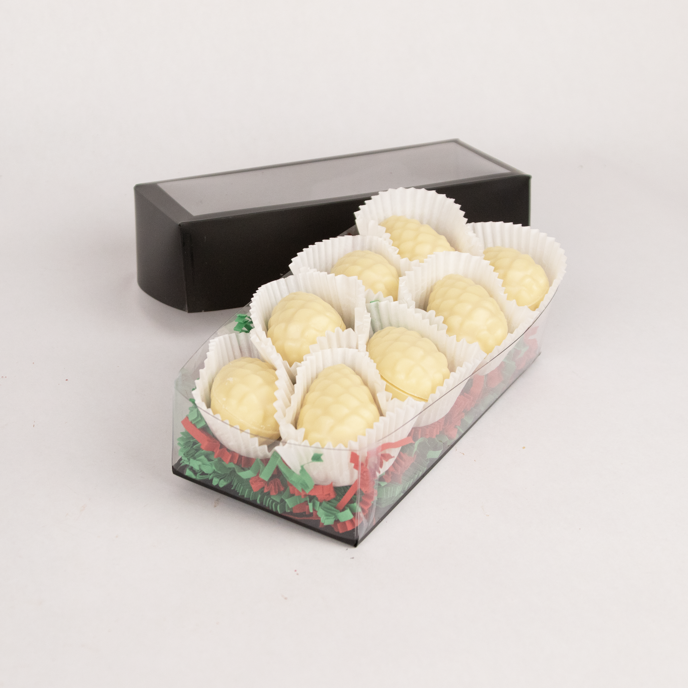 Clear gift box of eight Saratoga Chocolate Co. white chocolate pinecone-shaped praline bonbons, each nestled in white candy cups on colorful paper crinkle grass. The box features a clear lid with an elegant black border, showcasing the artisanal chocolate presentation. This product contains nuts.