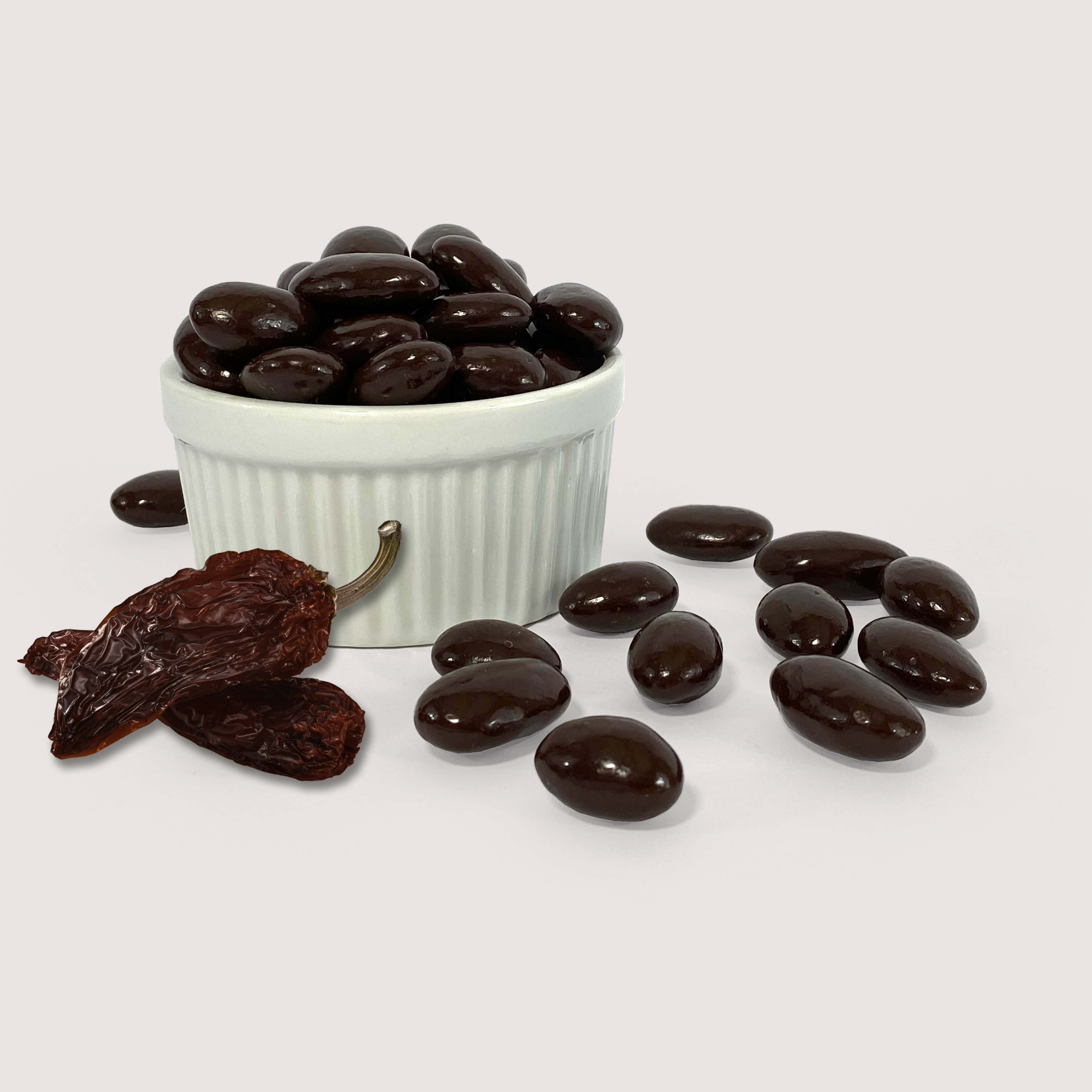 Image shows a pile of dark chocolate coated chipotle spiced almonds displayed in a fluted white bowl with some dark chocolate coated chipotle spiced almonds and two chipotle chilis scattered in front of the bowl.