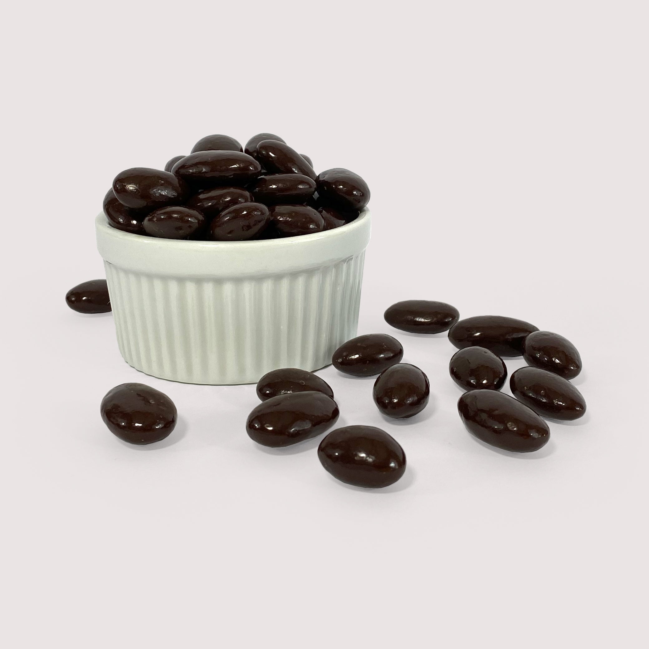 Pile of whole almonds coated in glossy dark chocolate, displayed in a white fluted bowl with several scattered in front.