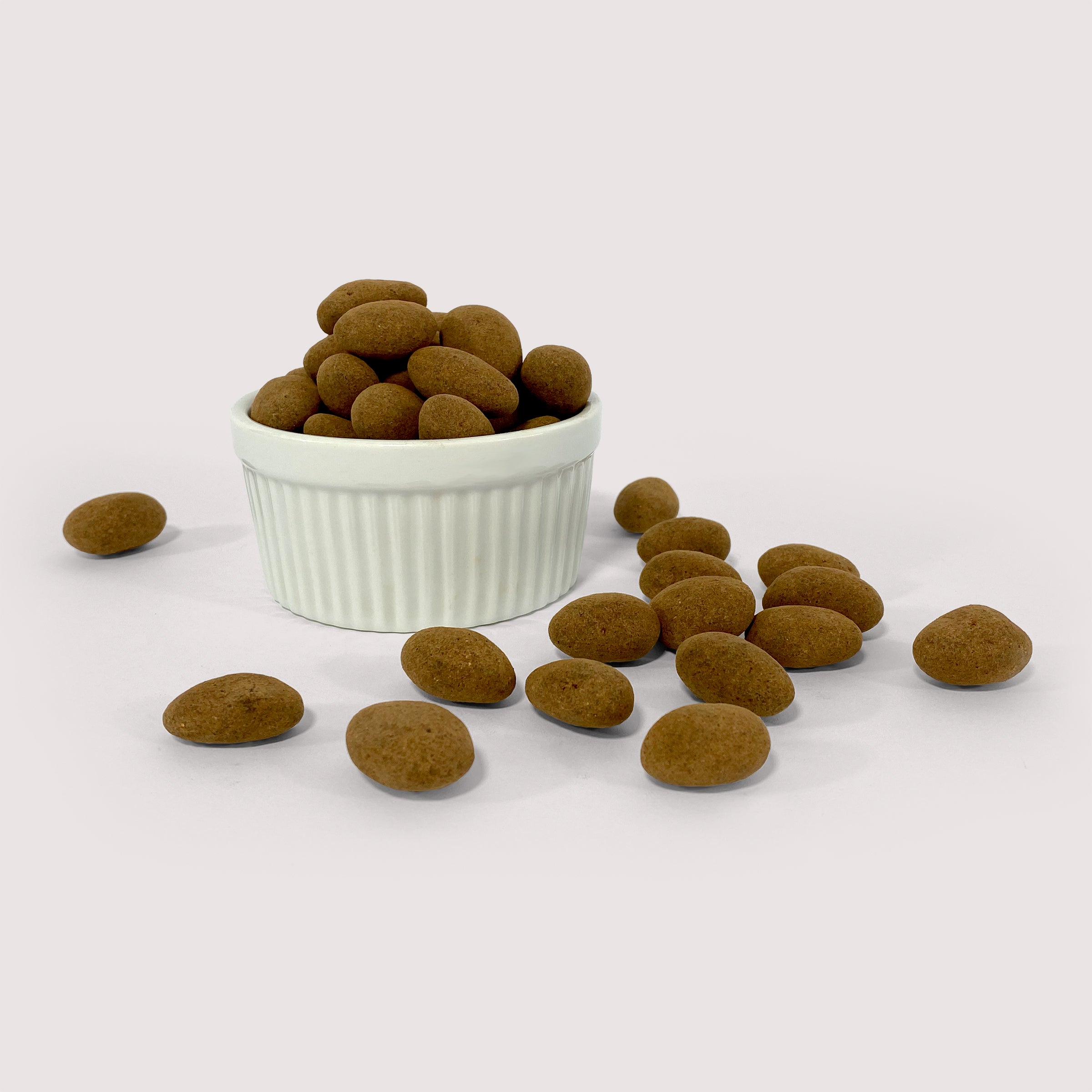 Image shows a pile of cocoa powder dusted chocolate Mexican vanilla spiced almonds displayed in a fluted white bowl with some cocoa powder dusted chocolate Mexican vanilla spiced almonds scattered in front 