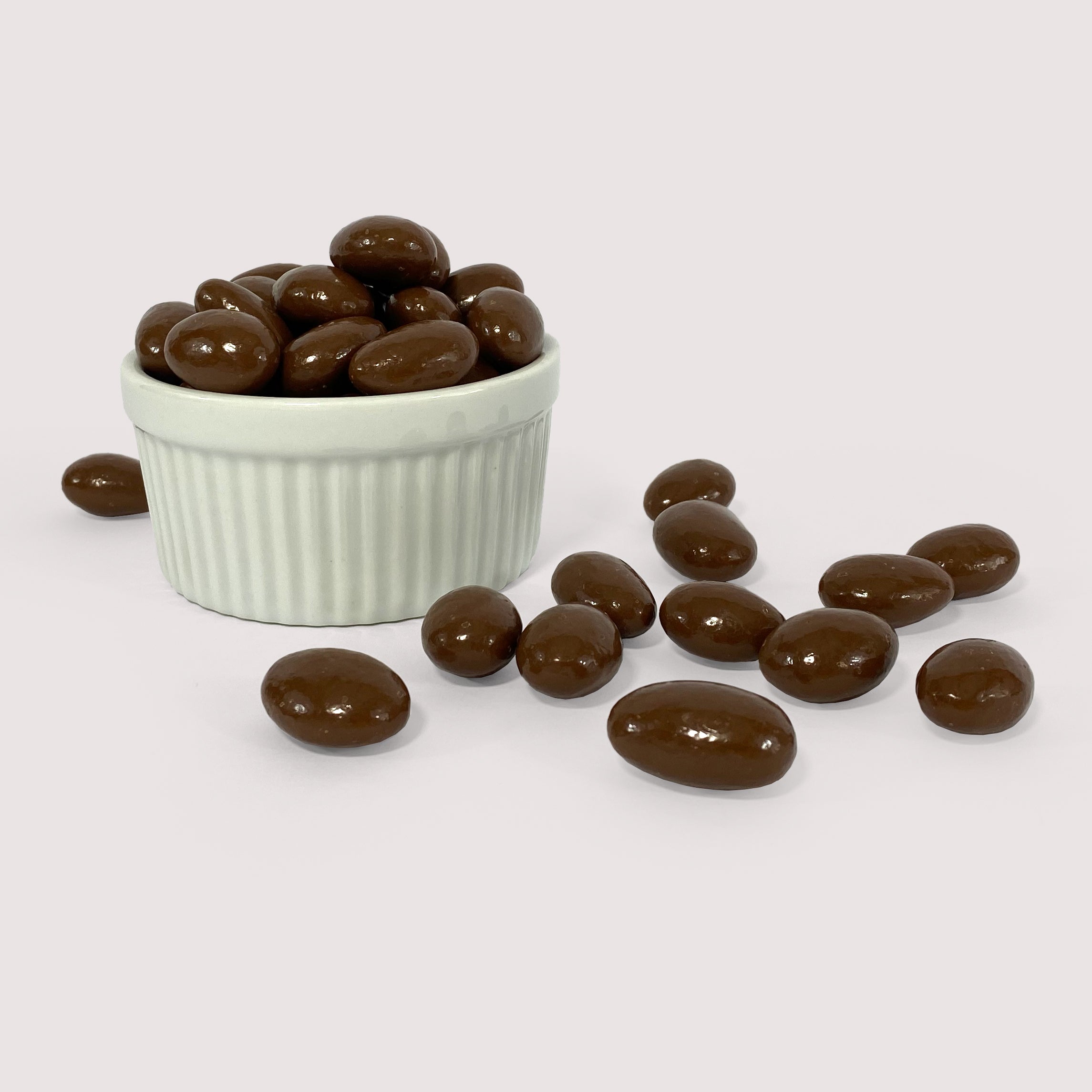Image shows a pile of milk chocolate coated sea salt almonds displayed in a fluted white bowl with pile of milk chocolate sea salt almonds scattered in front. 