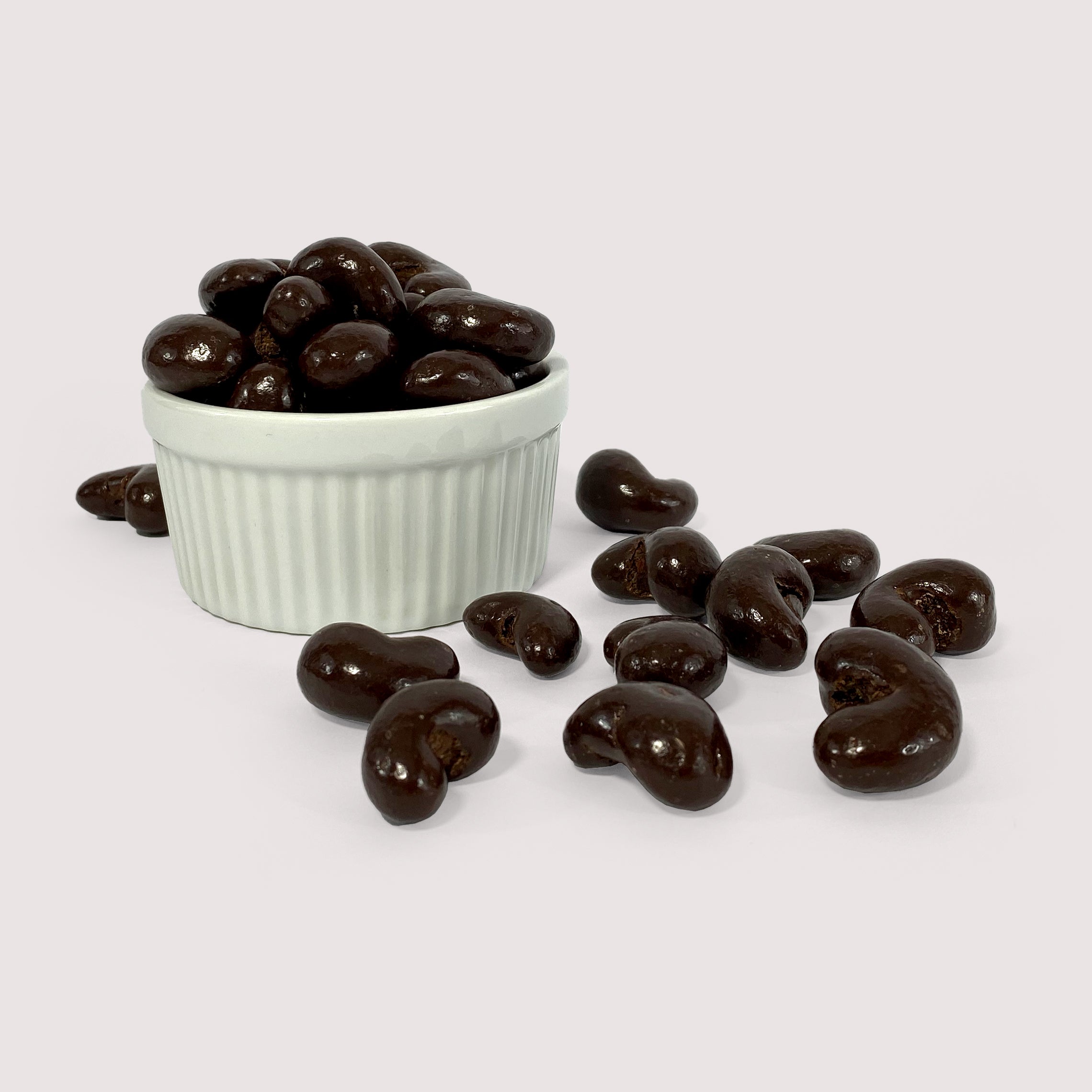 Image shows a pile of dark chocolate coated pecans displayed in a fluted white bowl with pile of dark chocolate coated pecans scattered in front.