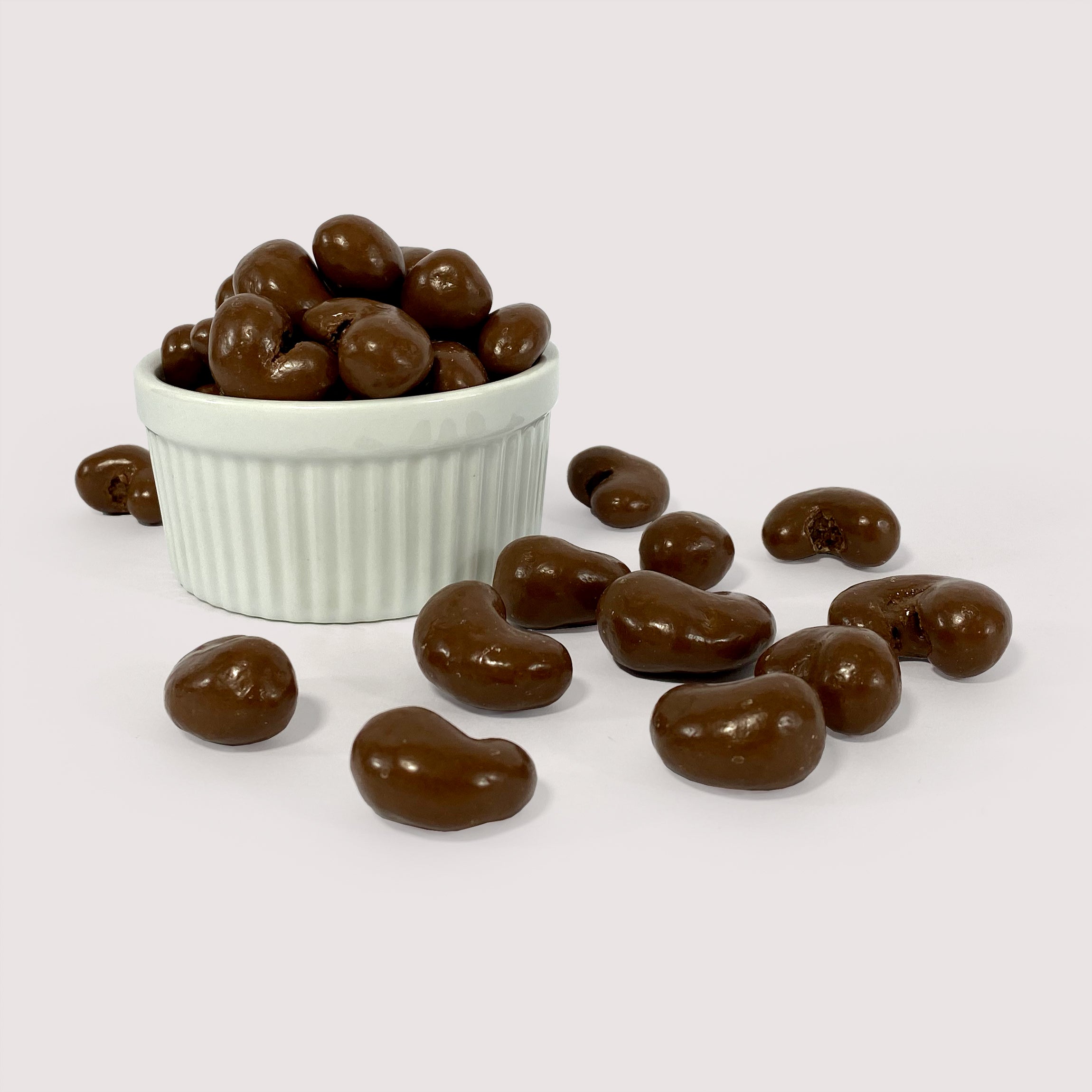 Image shows a pile of milk chocolate coated cashews displayed in a fluted white bowl with pile of milk chocolate coated cashews scattered in front. 